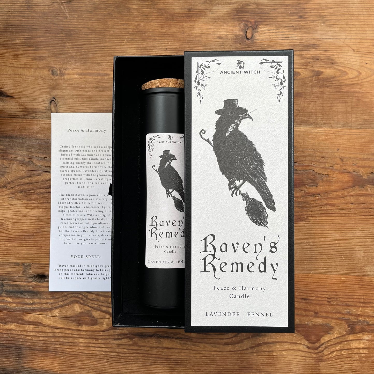 A black cylindrical candle in a black gift box with a cork top, shown beside its white printed outer sleeve and a small instruction card on a wooden surface. The packaging features a black raven illustration and the text 'Ancient Witch Raven's Remedy' and 'Lavender + Fennel'.