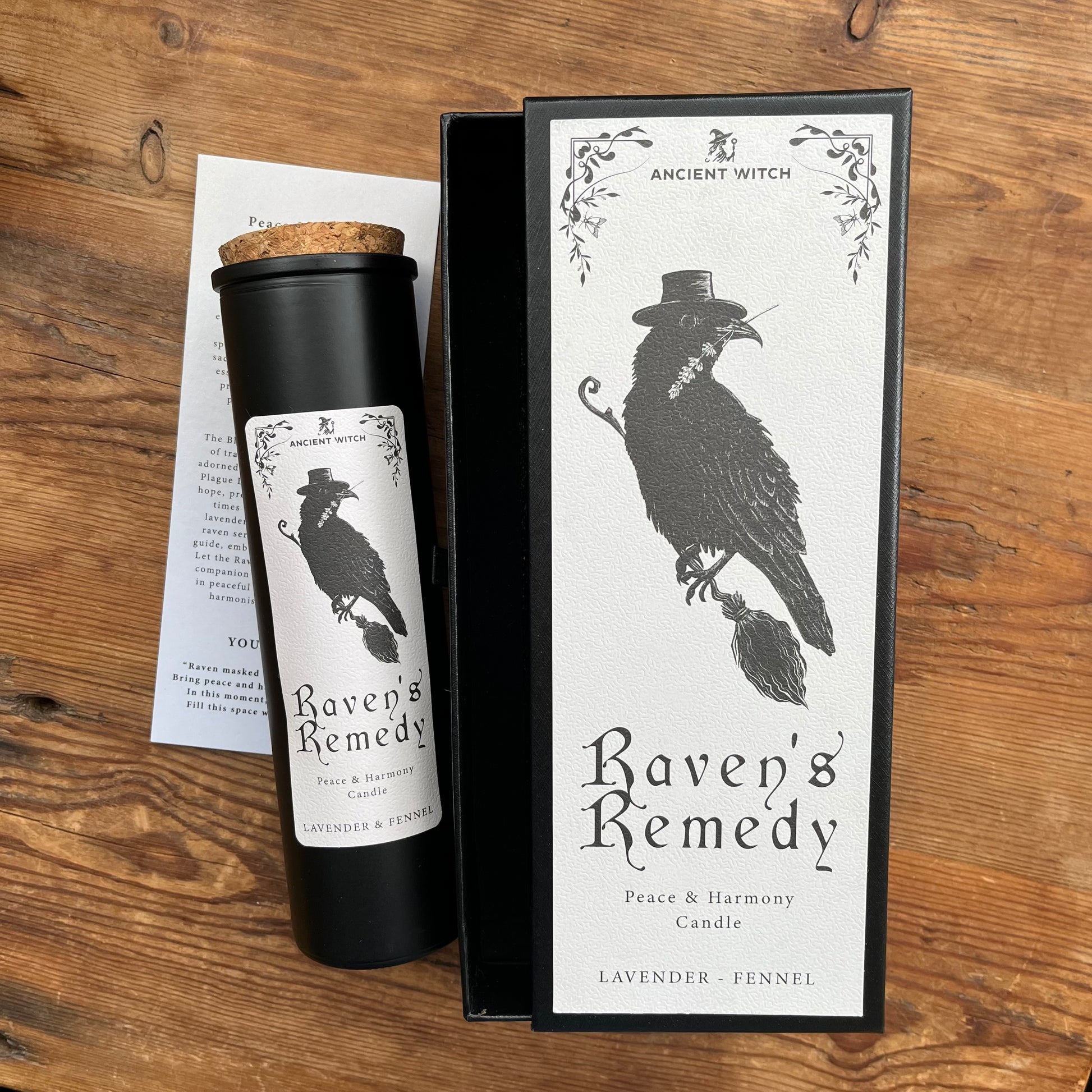 A black cylindrical candle jar with a cork stopper and a white label reading 'Ancient Witch Raven's Remedy' sits beside its black presentation box on a wooden surface. The box features a large black raven illustration and the same product name.