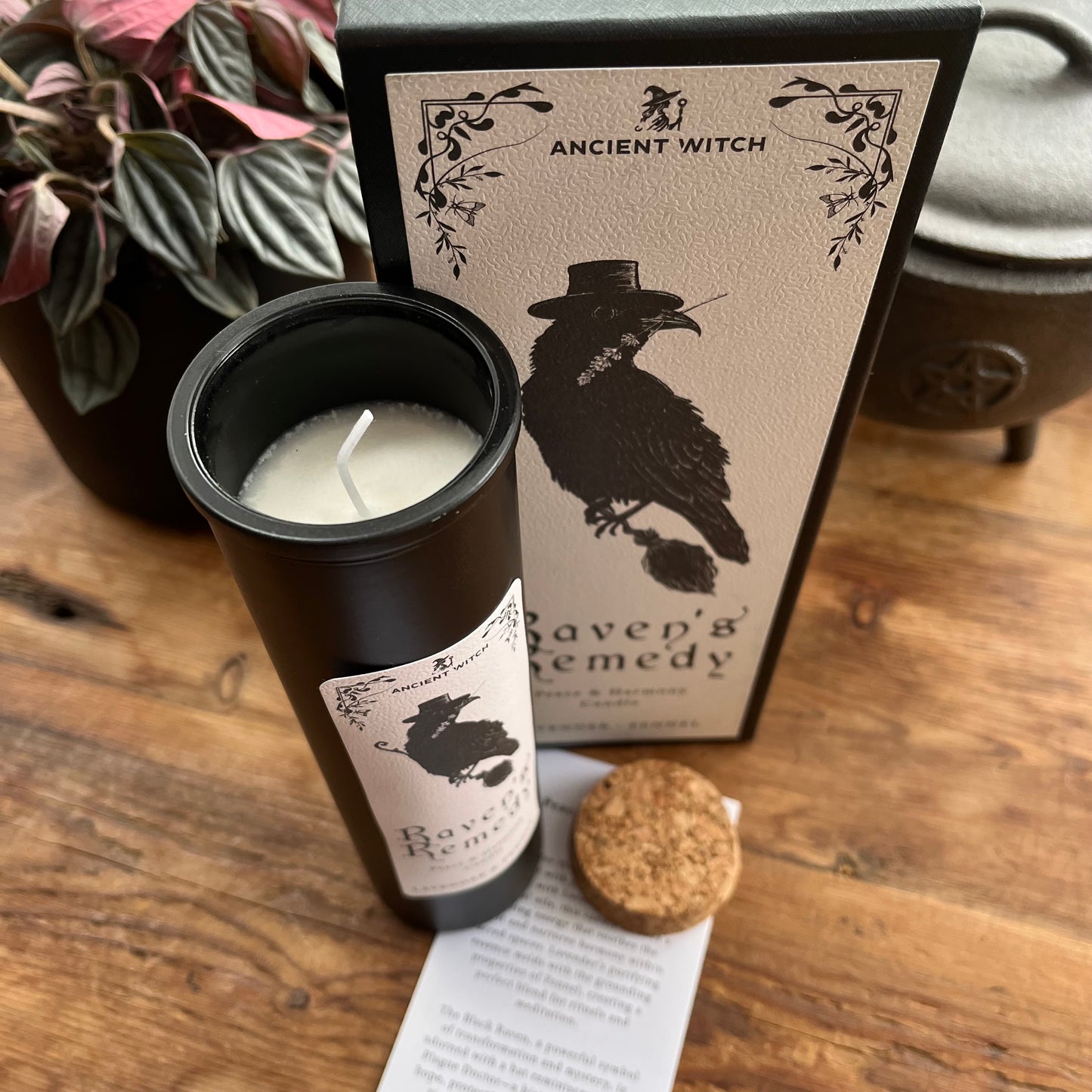 A black candle jar labeled 'Raven's Remedy' by Ancient Witch, shown with its matching product box, a cork stopper, and a small instruction card on a wooden tabletop.
