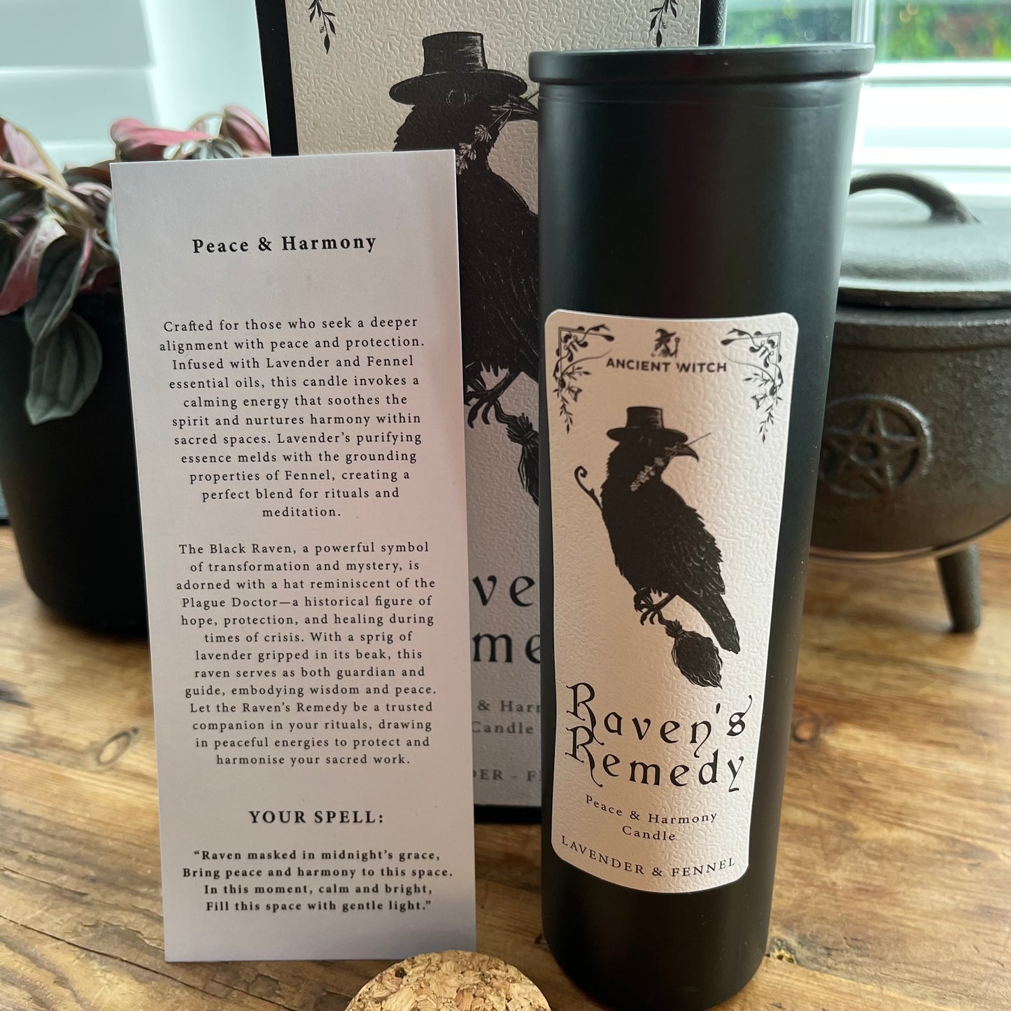 A black cylinder candle with a white 'Raven's Remedy' label stands next to a printed card titled 'Peace & Harmony' on a tabletop near a window.