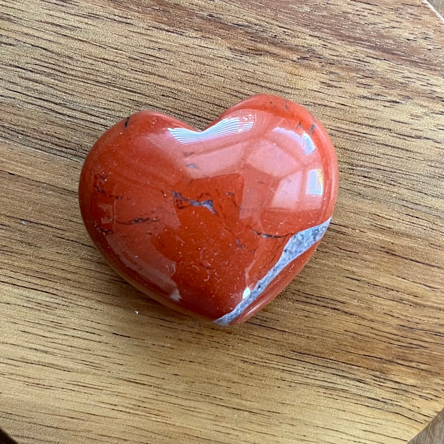 A polished heart-shaped red jasper stone with natural inclusions, resting on a light brown wooden surface.
