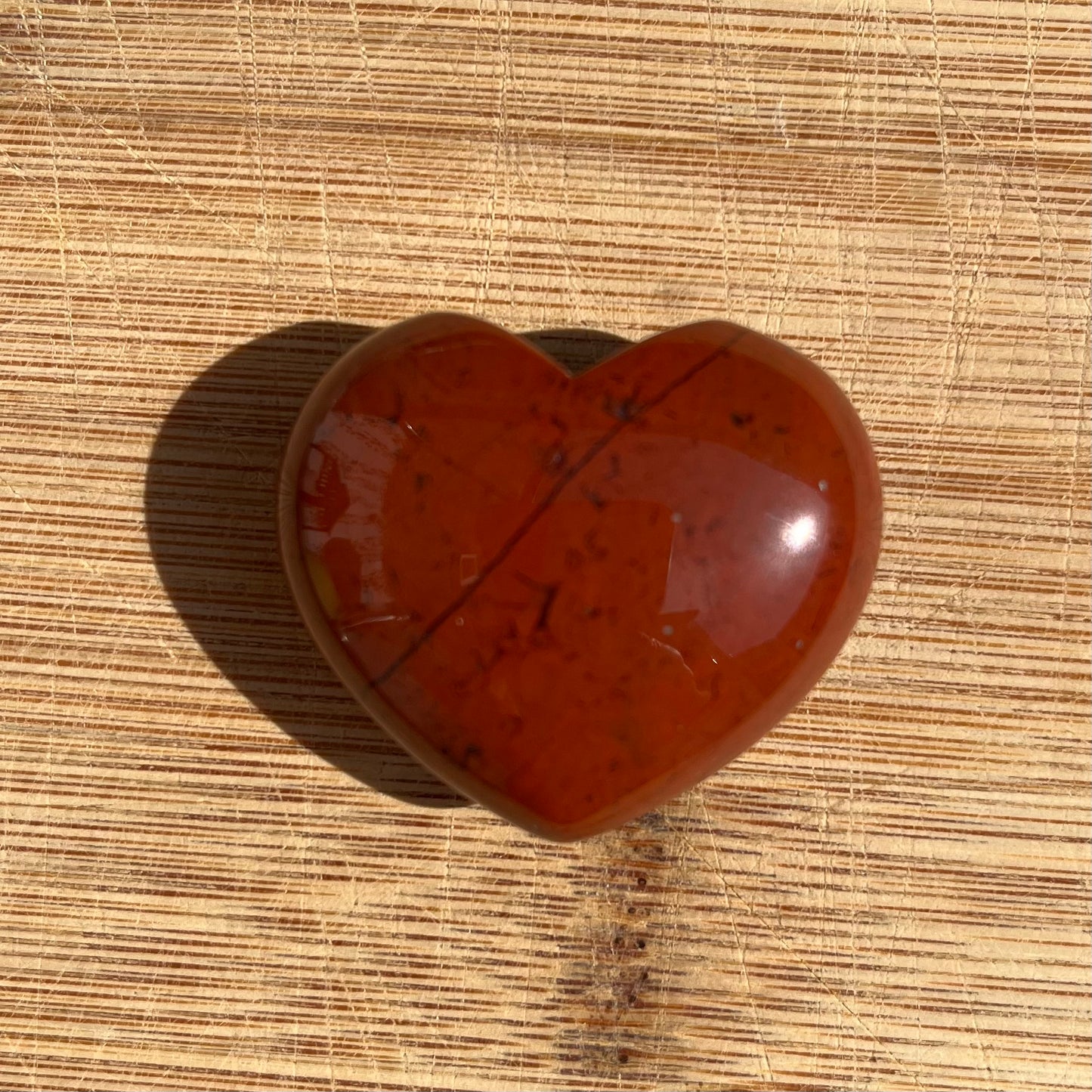 A heart-shaped polished red jasper stone with natural inclusions, resting on a light brown wood grain surface.
