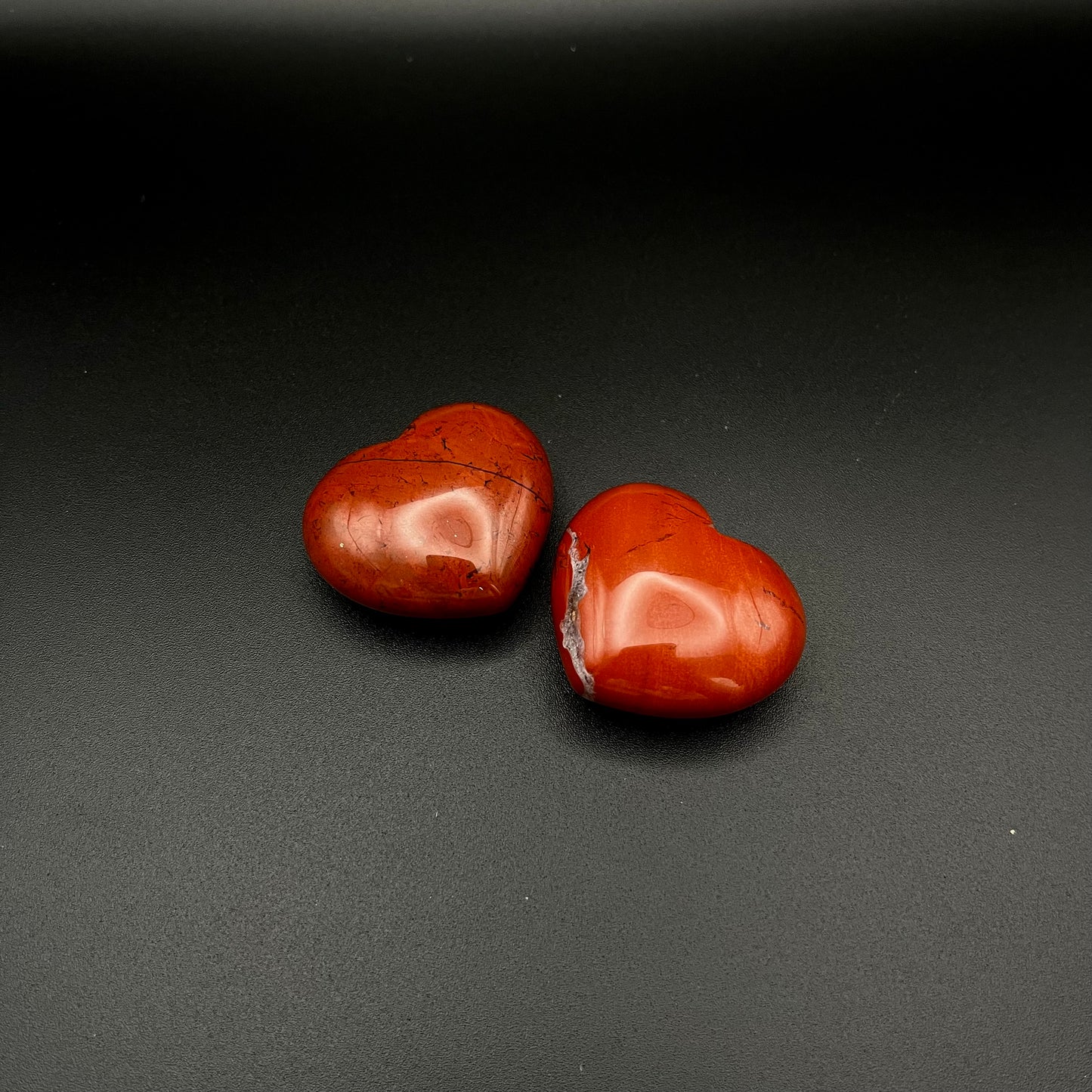 Two polished red heart-shaped stones, tumbled Red Jasper, displayed on a dark textured background.