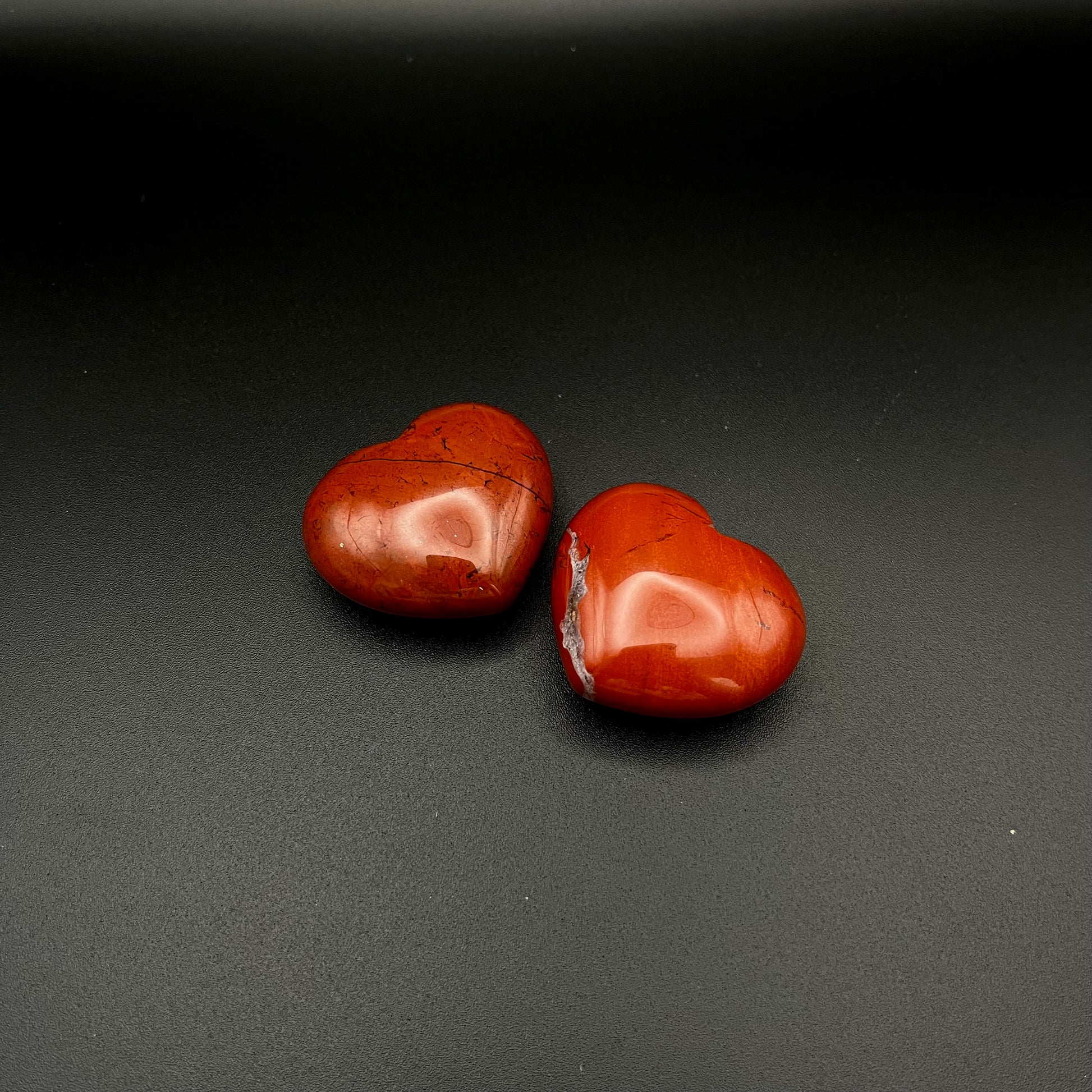 Two polished red heart-shaped stones, tumbled Red Jasper, displayed on a dark textured background.