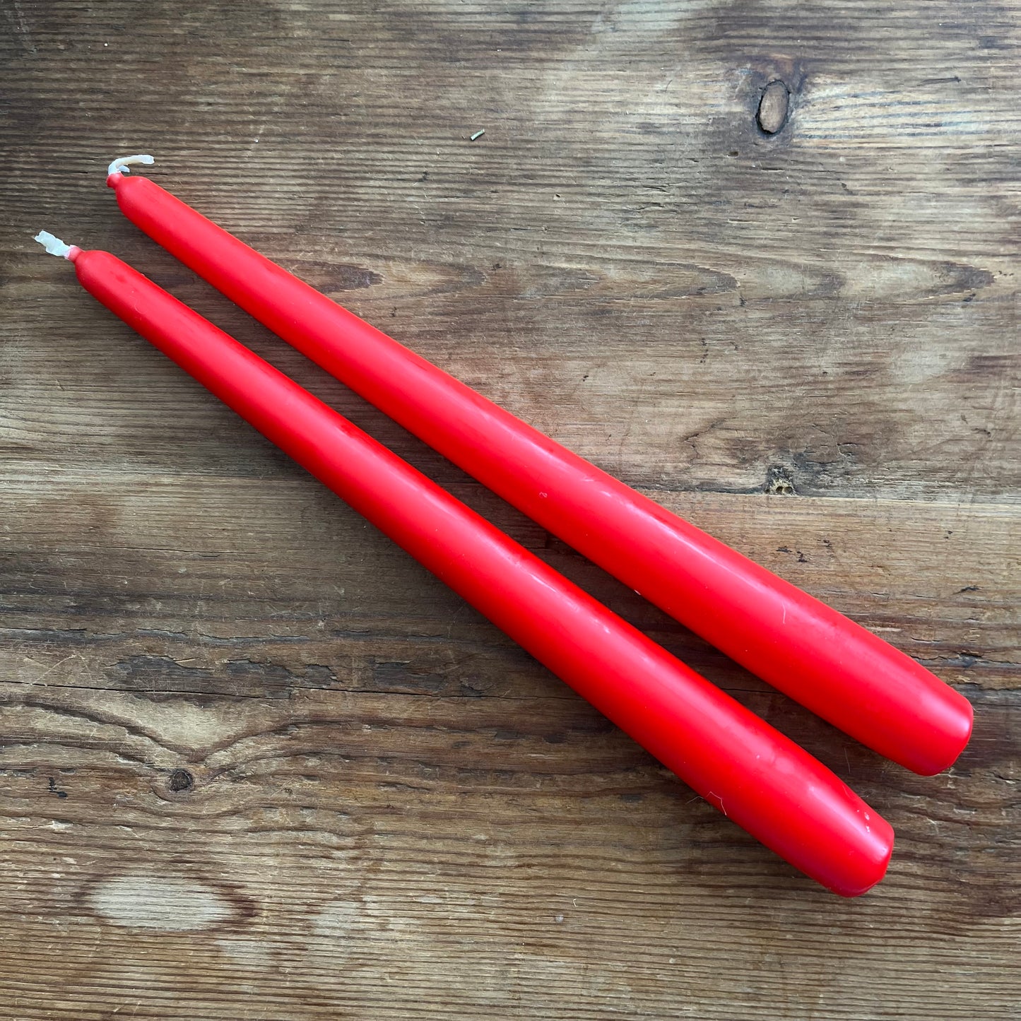 Two red taper candles with white wicks, placed diagonally on a rustic wooden surface.