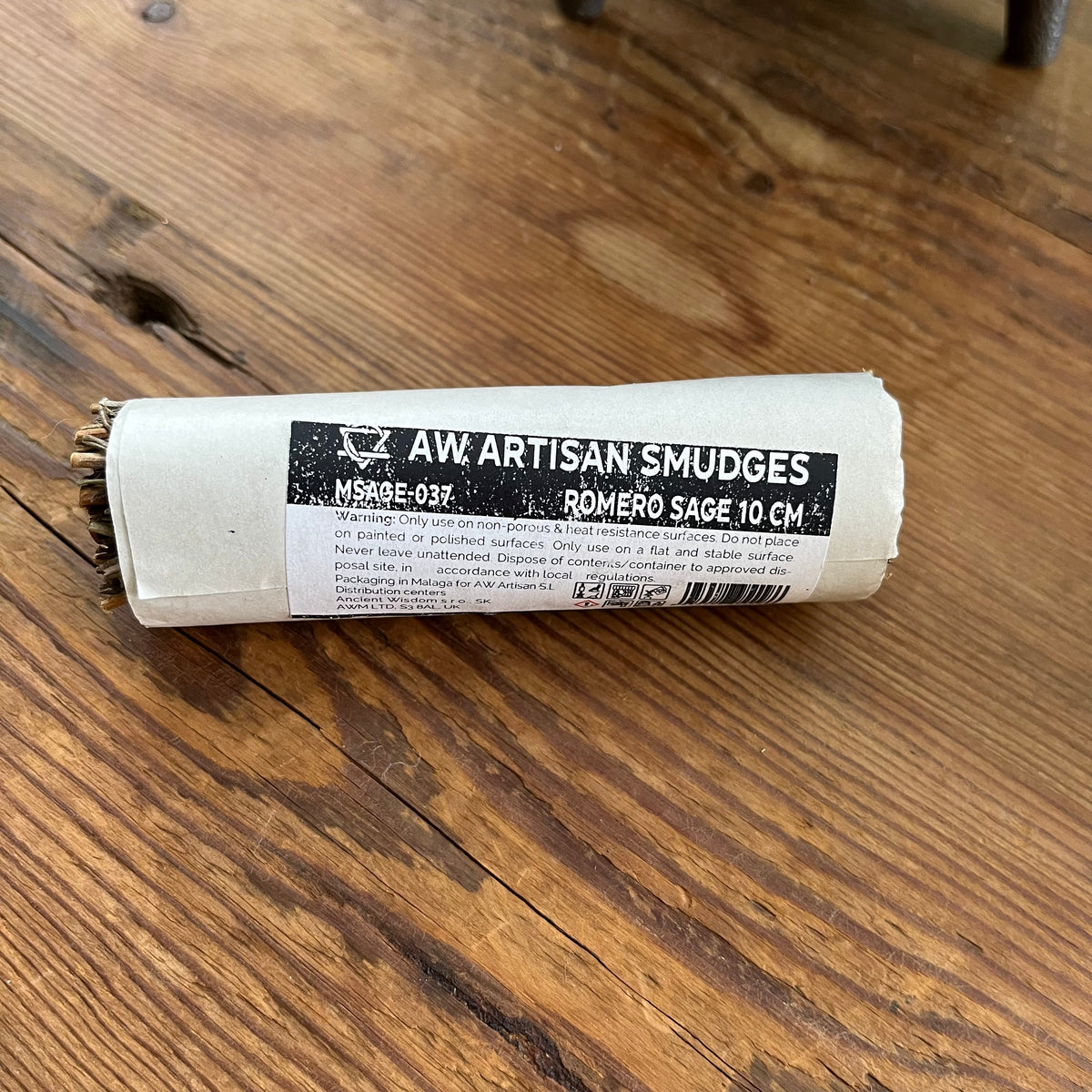 A roll of AW Artisan Smudges Romero Sage, 10 cm, wrapped in a white paper sleeve with a black label, resting on a rustic wooden table.