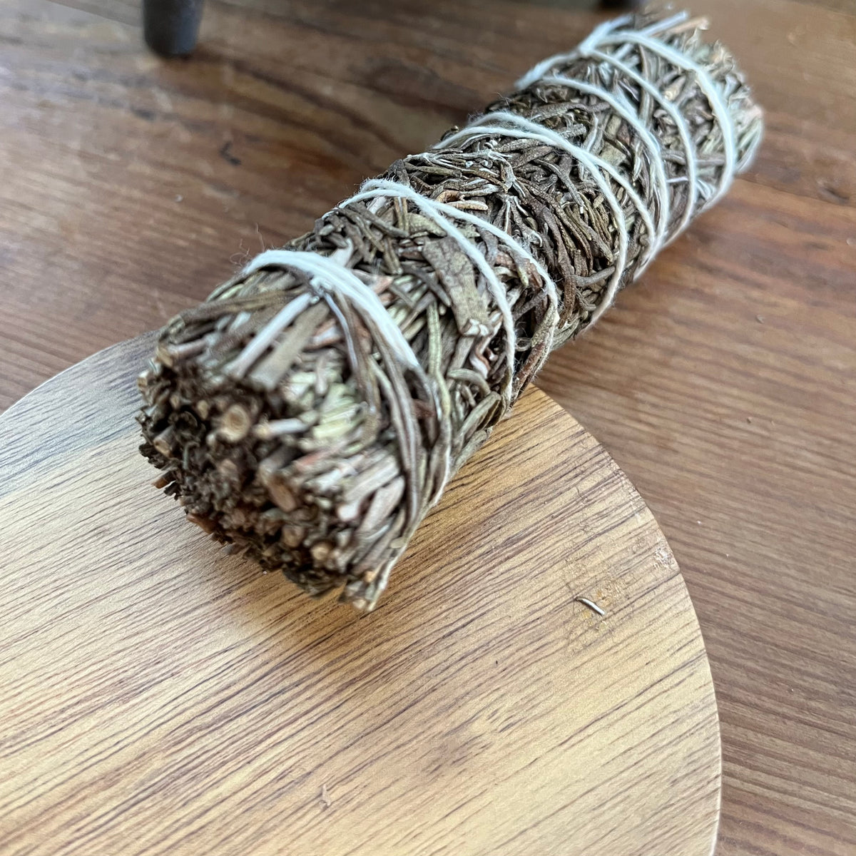 A dried herb smudge bundle wrapped in white string, resting diagonally on a round wooden coaster over a wooden tabletop.