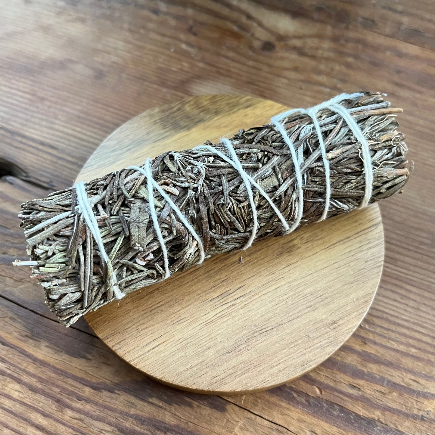 A dried herb smudge bundle wrapped in white string, resting diagonally on a round wooden coaster over a rustic wooden tabletop.