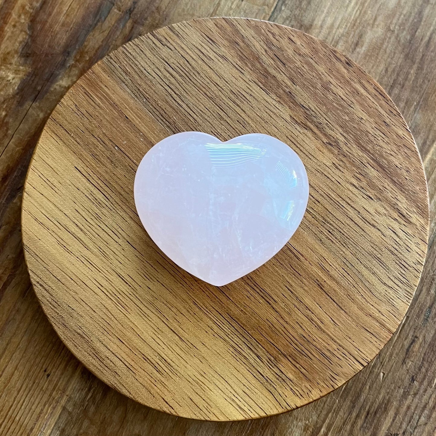 A heart-shaped pink stone, a rose quartz crystal, resting on a round wooden coaster over a wooden table.