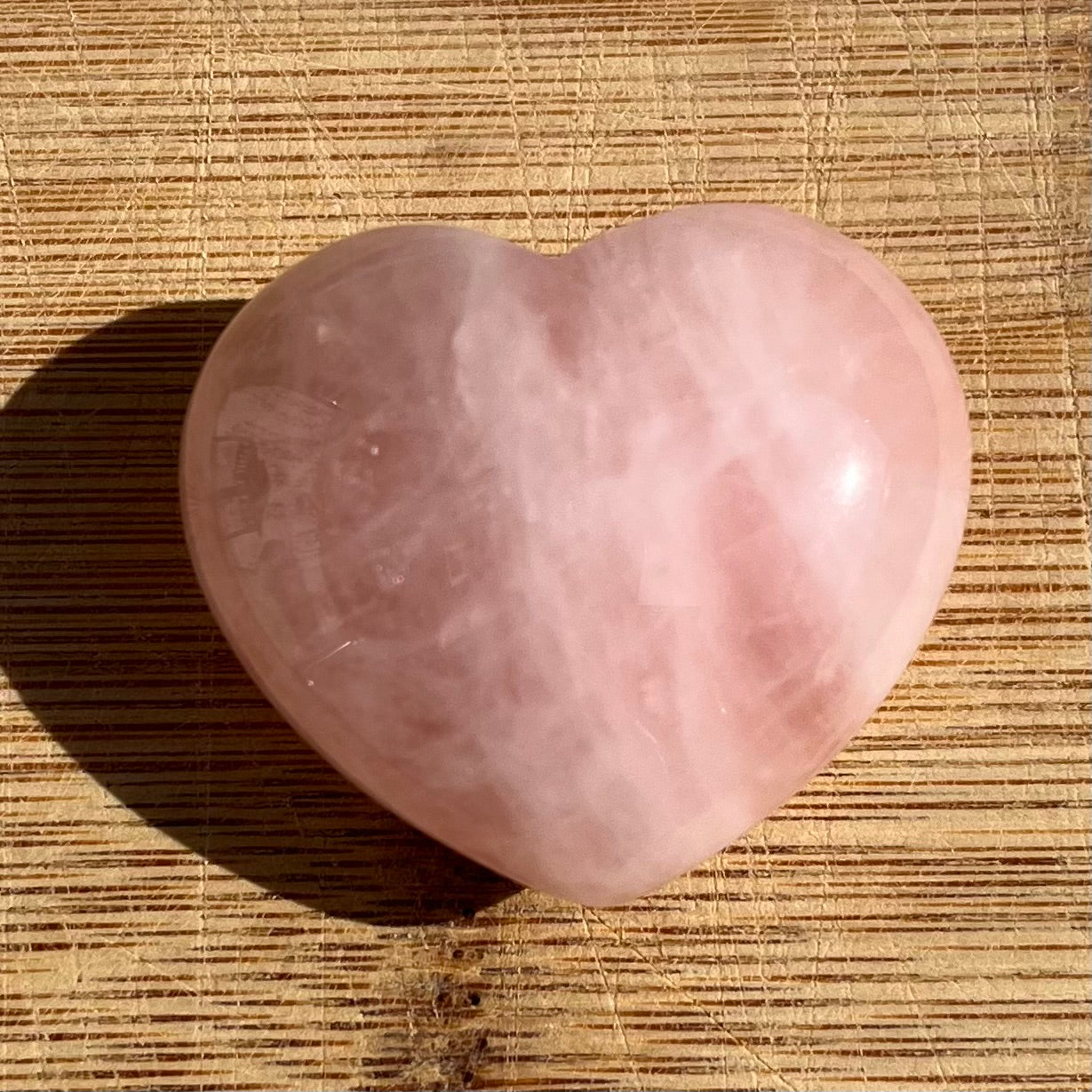 A polished pink heart-shaped rose quartz crystal resting on a textured bamboo mat, casting a soft shadow to the left.