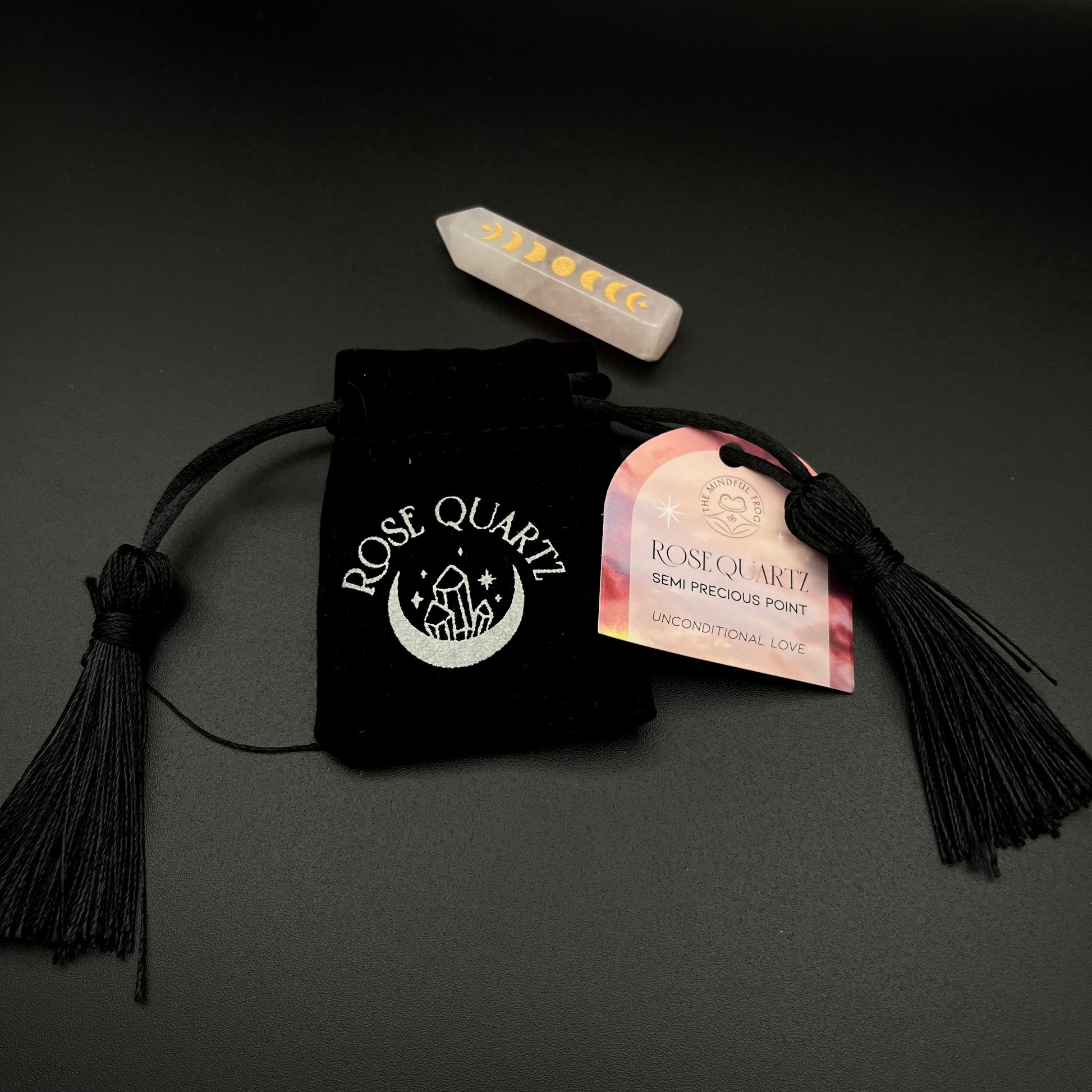 Rose Quartz semi-precious point crystal with a black velvet pouch, black tassels, and a pink and gold hangtag on a dark gray background.