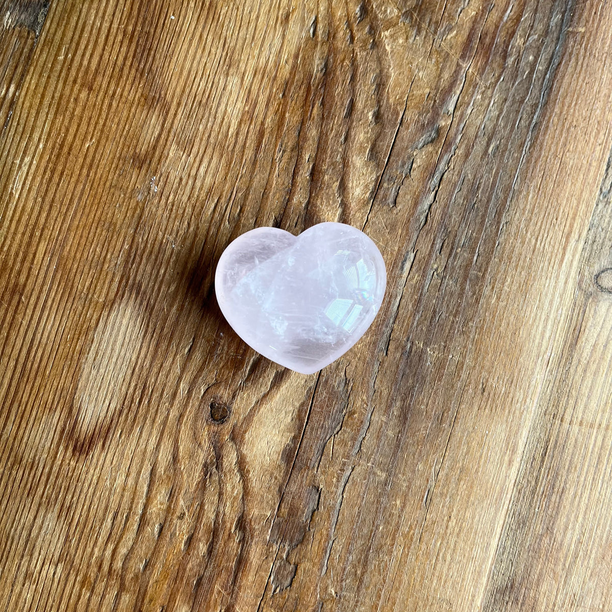 A small polished heart-shaped pink stone, likely rose quartz, resting on a rustic brown wooden surface.