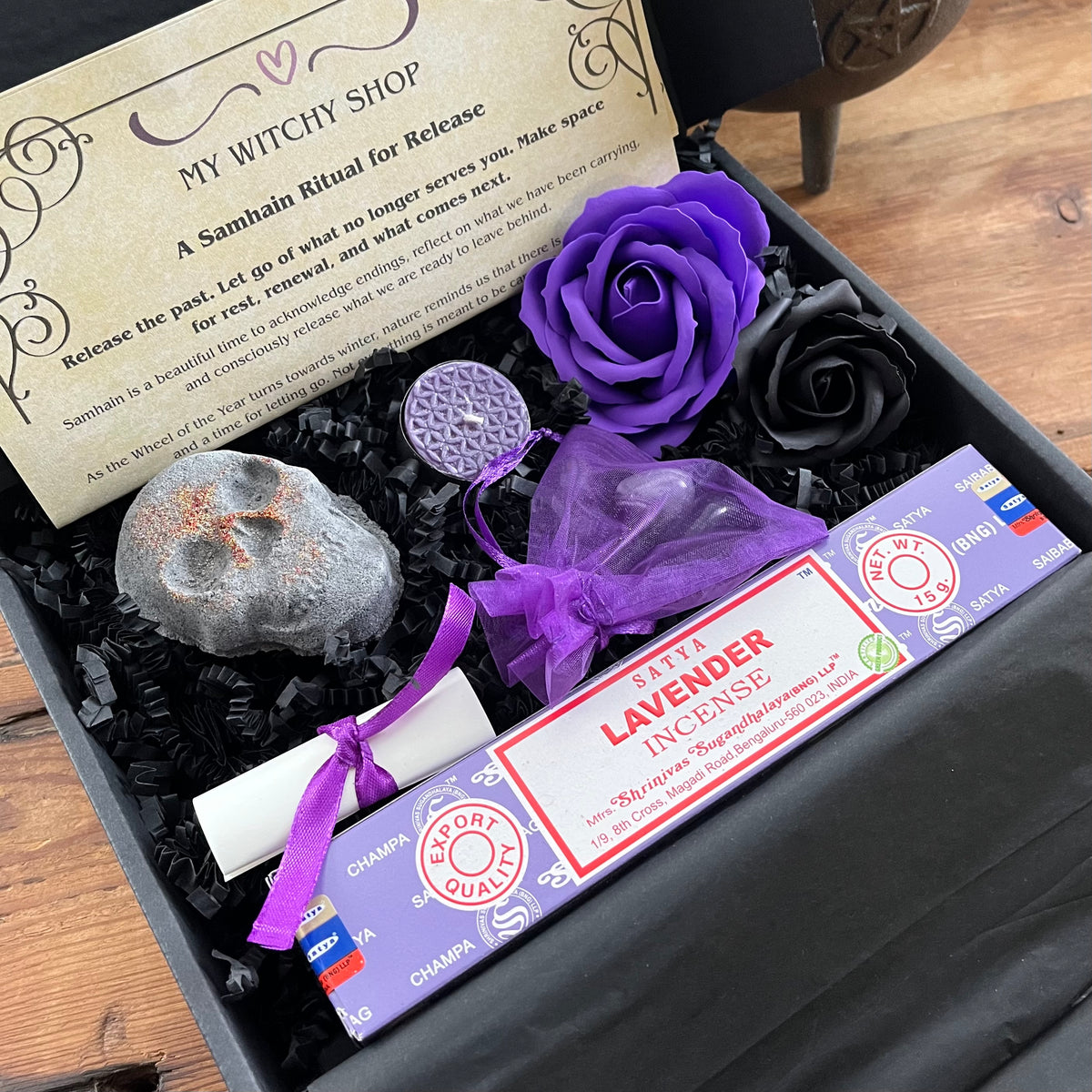 A black gift box filled with black shredded paper contains a lavender incense stick package tied with a purple ribbon, a small white scroll, a skull shaped bath bomb, a purple rose and black rose craft soap, and a purple organza bag containing crystals . An information sheet reading 'MY WITCHY SHOP' is placed beside the contents on a wooden surface.