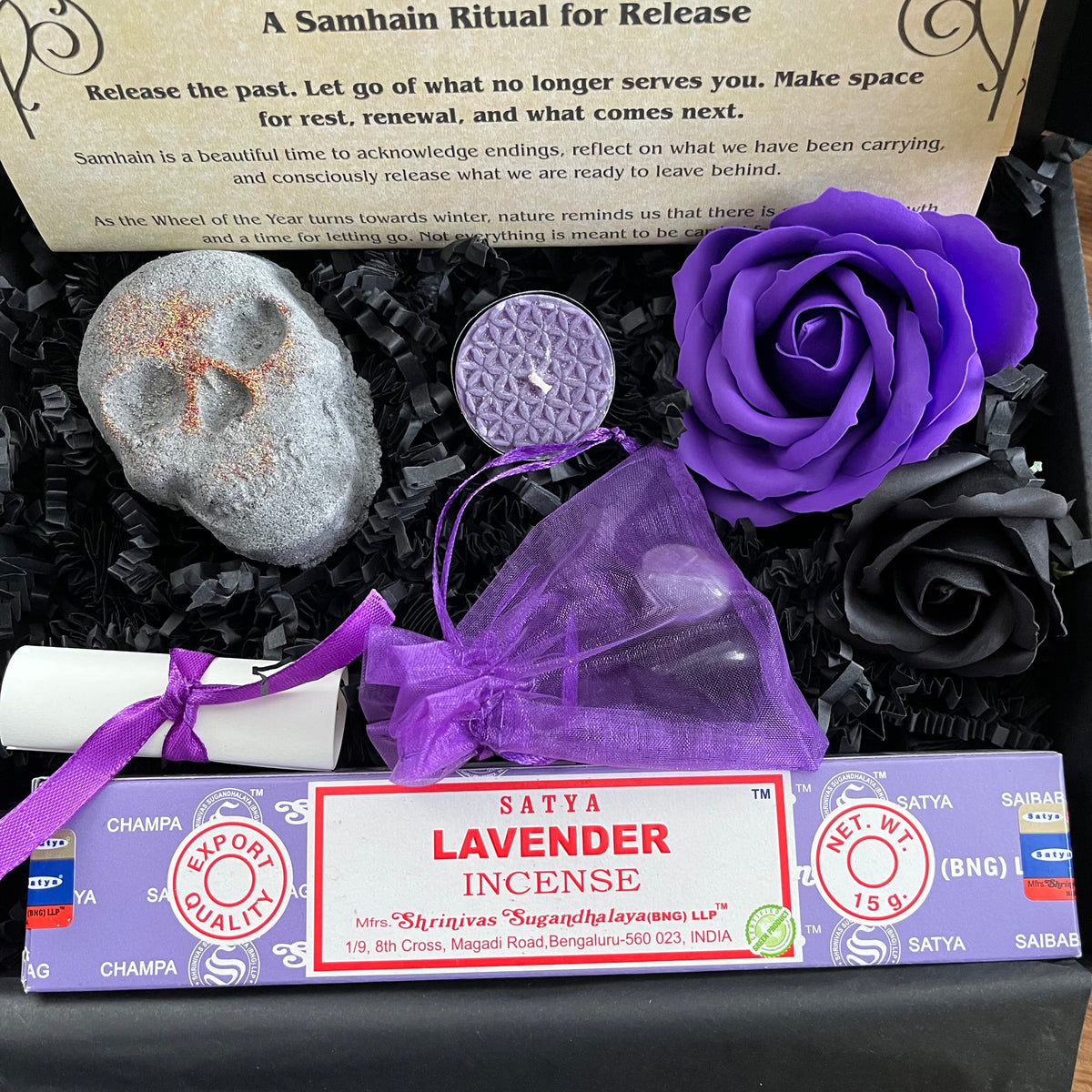 A boxed spiritual gift set featuring a lavender incense stick, a purple rose, a skull shaped bath bomb, a small purple pouch, and a printed information sheet with Sanskrit ritual text, arranged in black gift packaging.