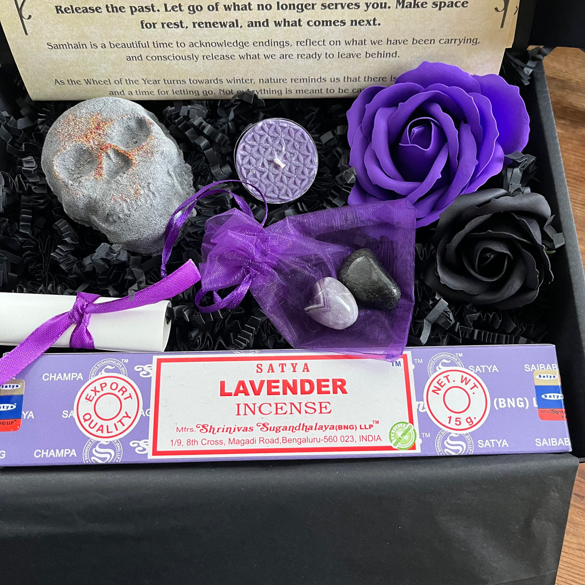 A black gift box filled with black shredded paper contains a lavender incense stick package tied with a purple ribbon, a small white scroll, a skull shaped bath bomb, a purple rose and black rose craft soap, and a purple organza bag containing crystals . An information sheet reading 'MY WITCHY SHOP' is placed beside the contents on a wooden surface.