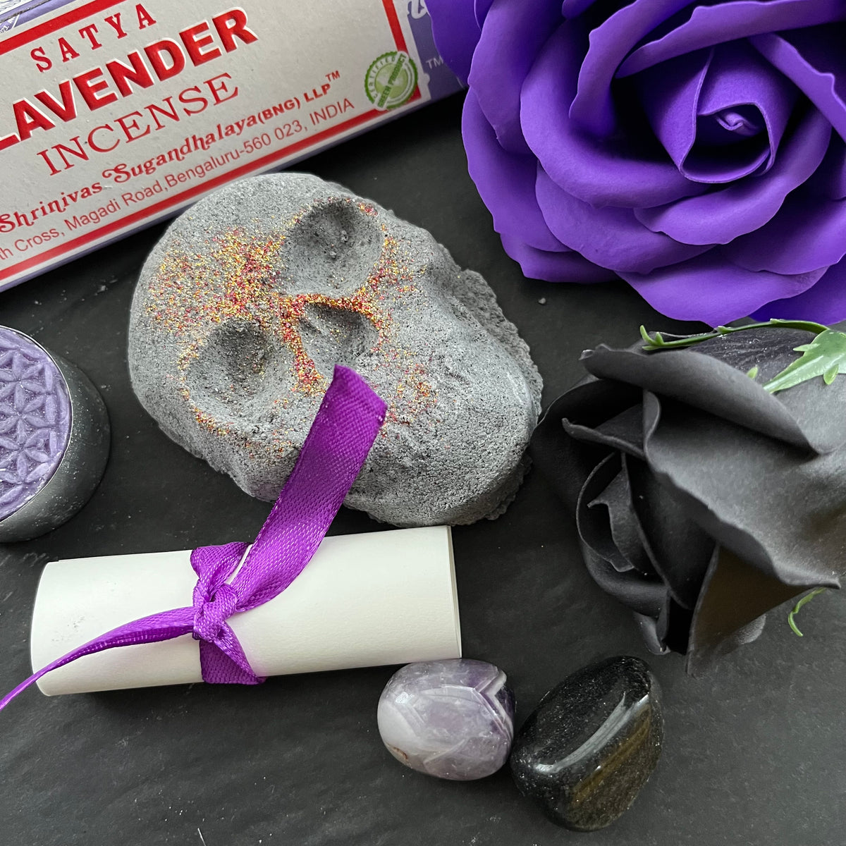 lavender incense sticks, package tied with a purple ribbon, a small white scroll, a skull shaped bath bomb, a purple rose and black rose craft soap, a purple candle and a purple organza bag containing crystals . An information sheet reading 'MY WITCHY SHOP' is placed beside the contents on a wooden surface.