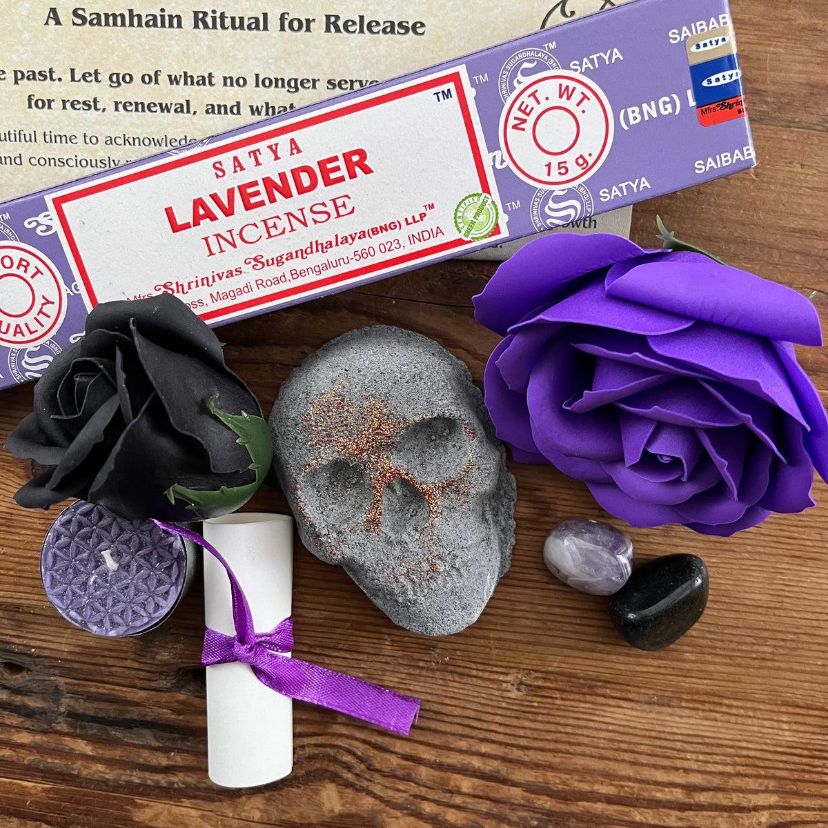 lavender incense sticks, package tied with a purple ribbon, a small white scroll, a skull shaped bath bomb, a purple rose and black rose craft soap, a purple candle and a purple organza bag containing crystals . An information sheet reading 'MY WITCHY SHOP' is placed beside the contents on a wooden surface.