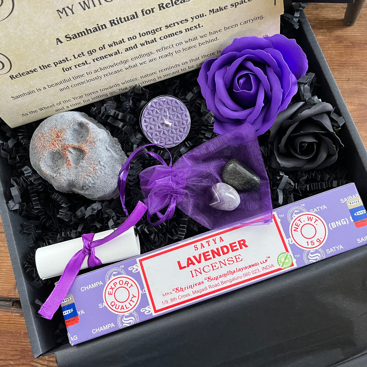 A black gift box filled with black shredded paper contains a lavender incense stick package tied with a purple ribbon, a small white scroll, a skull shaped bath bomb, a purple rose and black rose craft soap, and a purple organza bag containing crystals . An information sheet reading 'MY WITCHY SHOP' is placed beside the contents on a wooden surface.