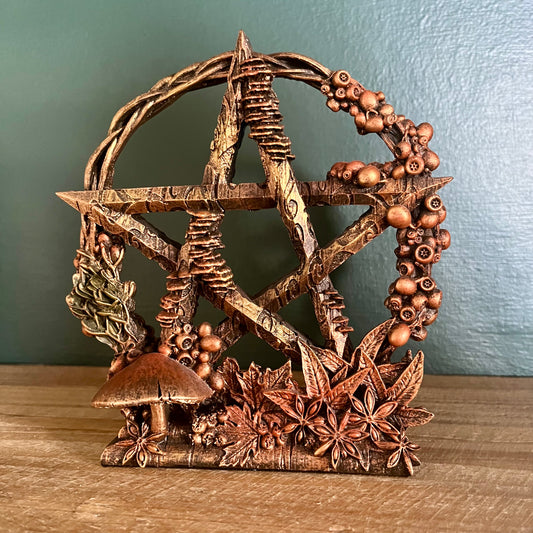 A rustic copper-colored metal pentacle star decoration with a woodland theme, featuring a mushroom, pine branches, acorns, and leaves, displayed on a wooden surface against a teal wall.
