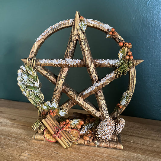 A decorative pentagram altar piece with a rustic weathered brown finish, adorned with faux snow, green pine branches, small orange berries, cinnamon sticks, and dried botanicals, displayed on a wooden surface against a dark teal wall.