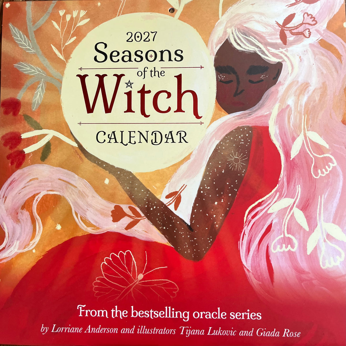 Front cover of the 2027 Seasons of the Witch Calendar, featuring an illustrated woman with long white hair, a red dress, and warm orange and pink background tones. The title text is prominently displayed in the center.