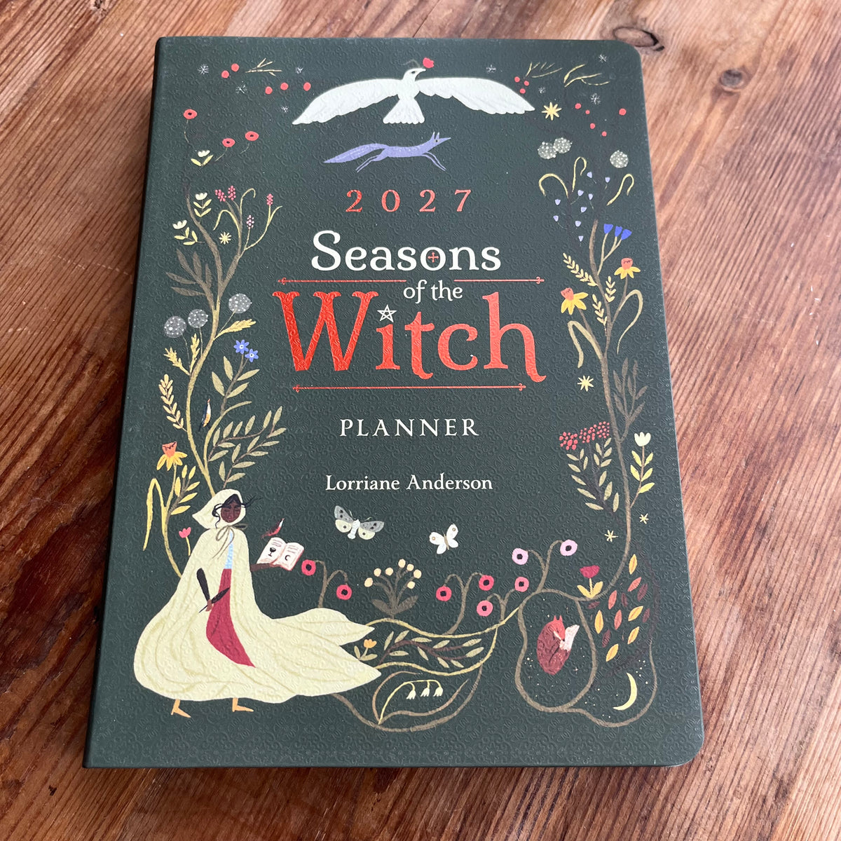A hardcover planner titled '2027 Seasons of the Witch PLANNER' by Lorriane Anderson, featuring a dark forest green cover decorated with whimsical witch illustrations and floral borders, placed on a wooden tabletop.