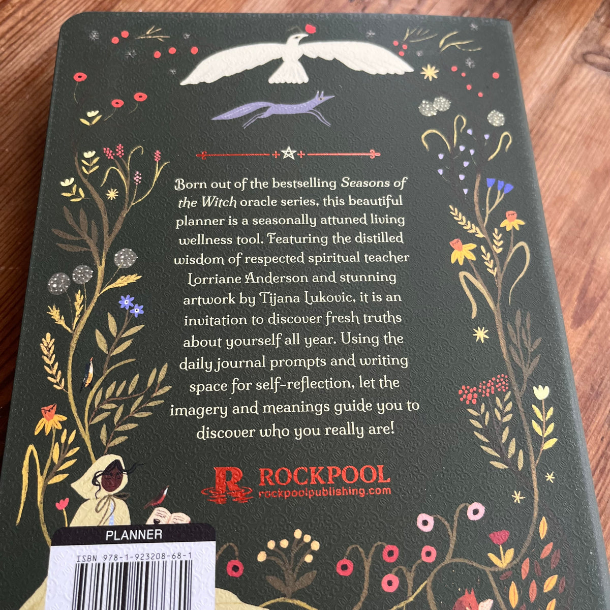 Back cover of a Rockpool journal or planner on a wooden surface, featuring a dark green background with whimsical floral illustrations and a white bird at the top. The text describes the journal's wellness and spiritual theme, with a barcode label at the bottom left.