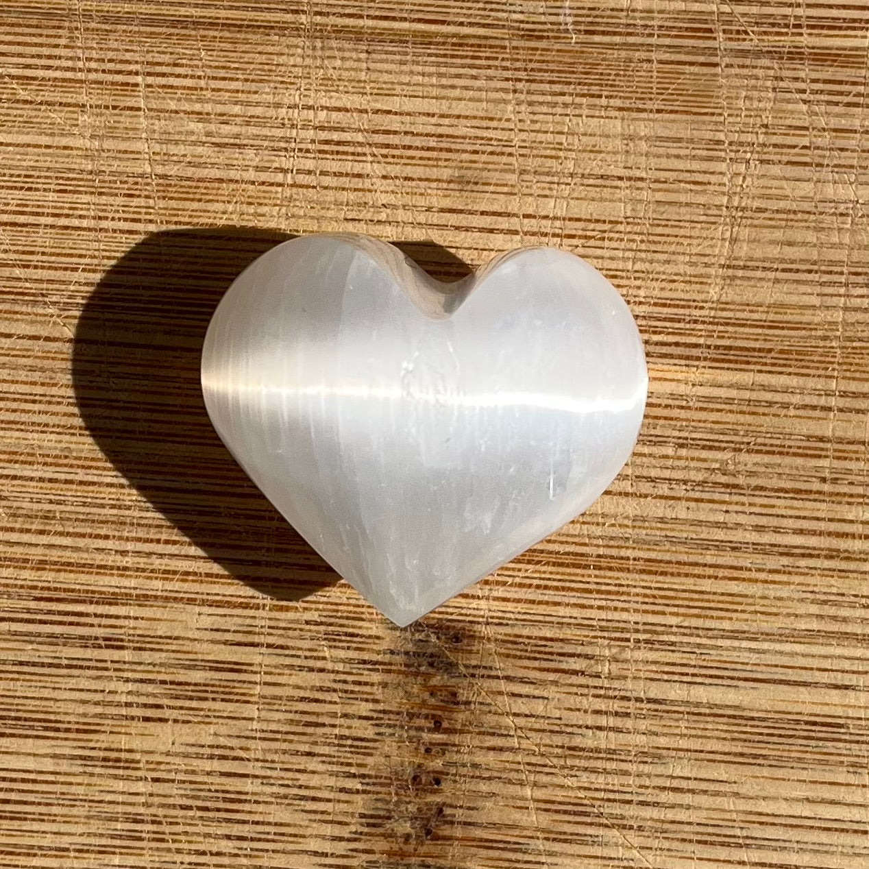 A heart-shaped Selenite stone with a polished, reflective surface, resting on a textured wooden surface.
