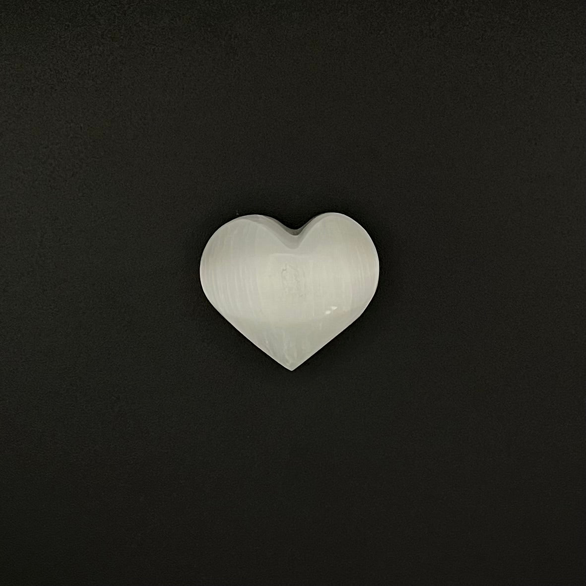 A small heart-shaped Selenite stone with a smooth, polished surface, centered on a solid black background.