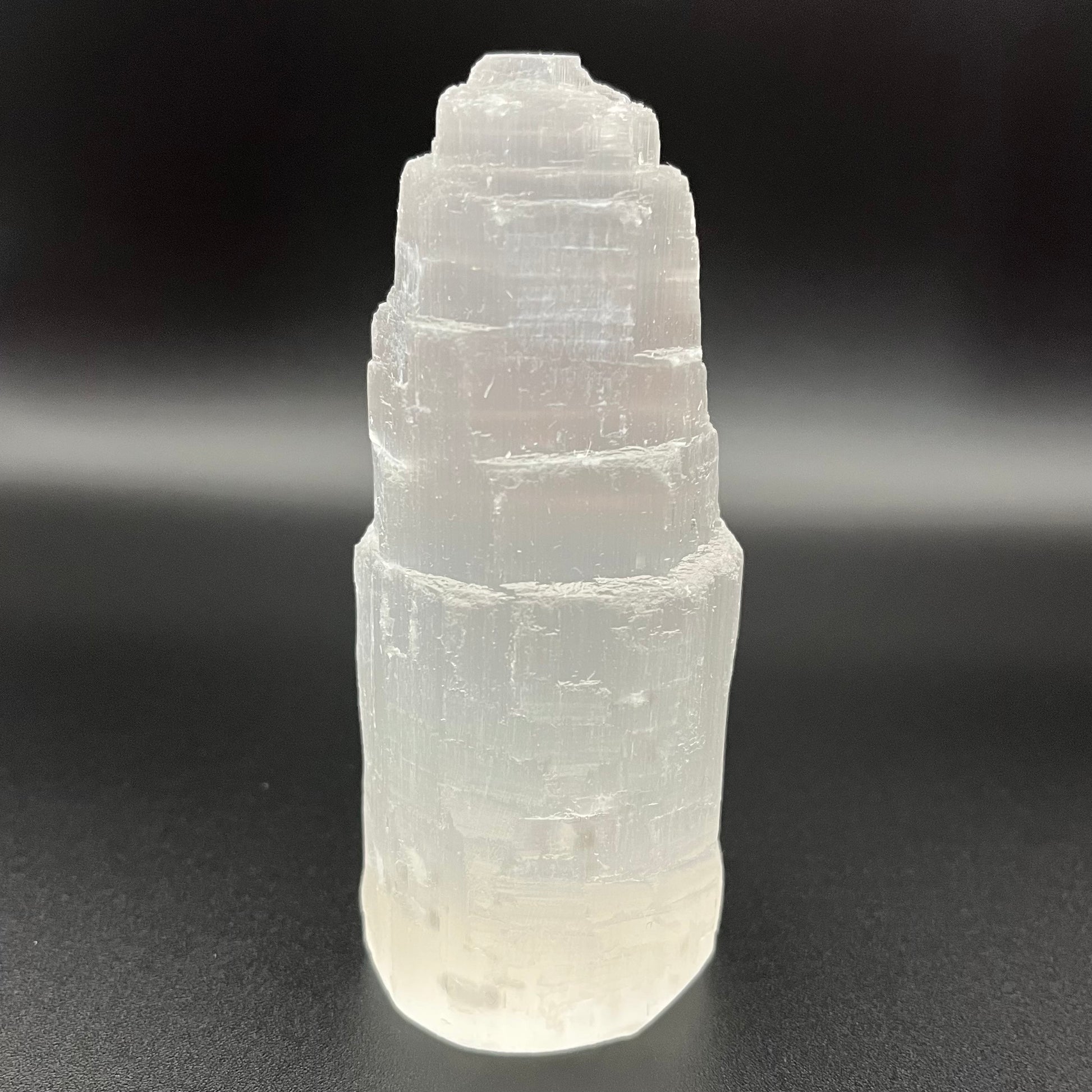 A natural selenite tower crystal with layered, stacked geometric facets, shown standing upright on a dark, neutral background.