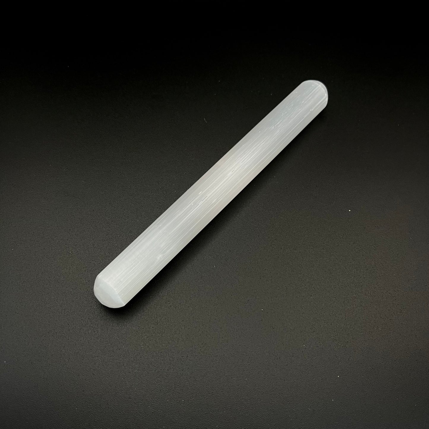 A translucent white cylindrical rod, a crystal wand or selenite stick, photographed diagonally on a dark textured background.