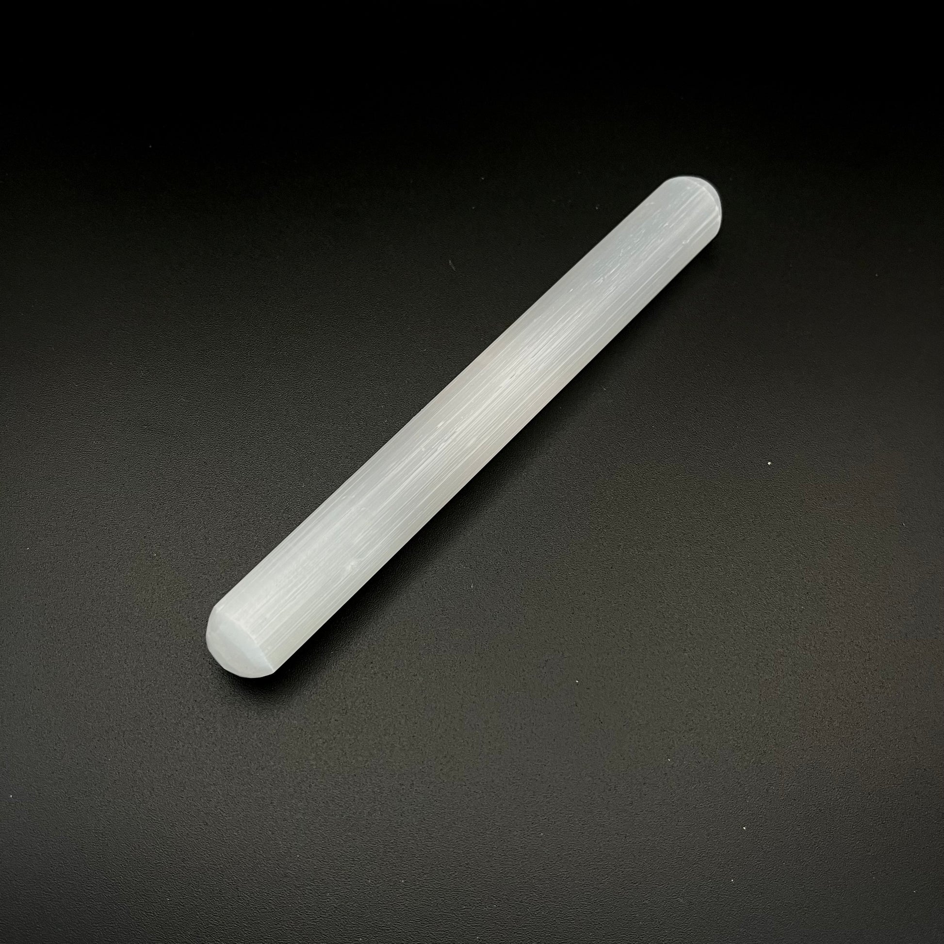 A translucent white cylindrical rod, a crystal wand or selenite stick, photographed diagonally on a dark textured background.