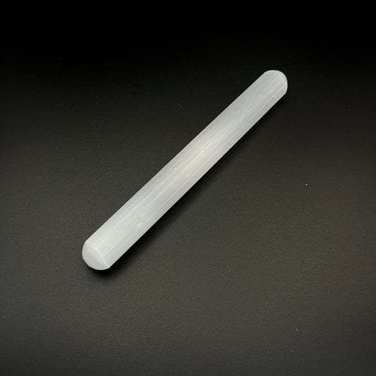 A translucent white cylindrical rod, a crystal wand or selenite stick, photographed diagonally on a dark textured background.