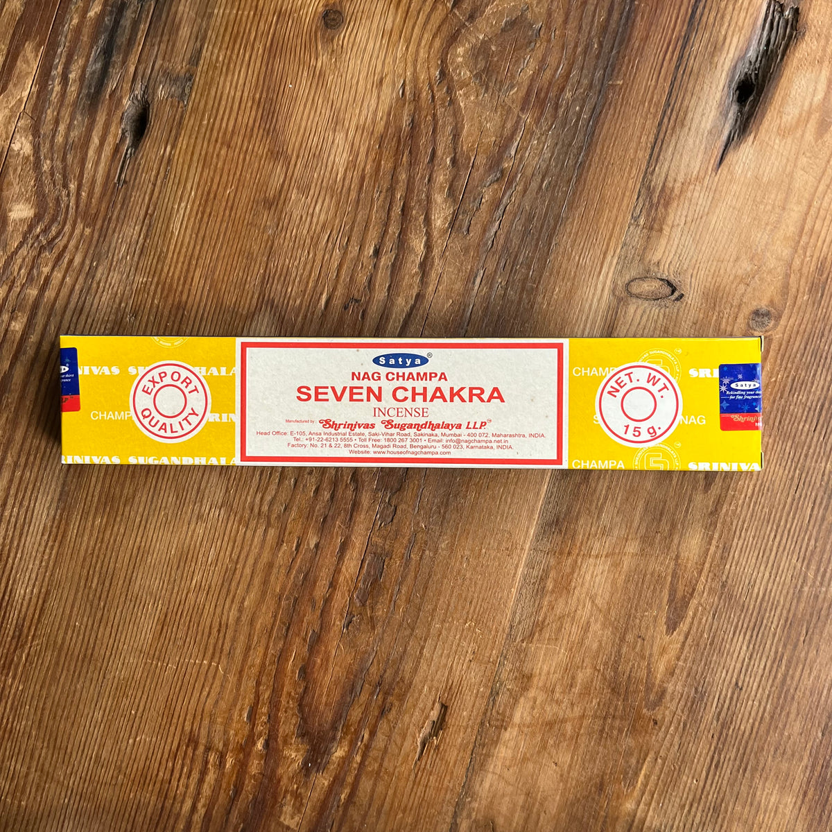 A rectangular box of Nag CHAMPA SEVEN CHAKRA INCENSE sticks lies on a rustic wooden surface. The packaging is primarily yellow with a central cream-colored label bordered in red, featuring blue and red brand details.