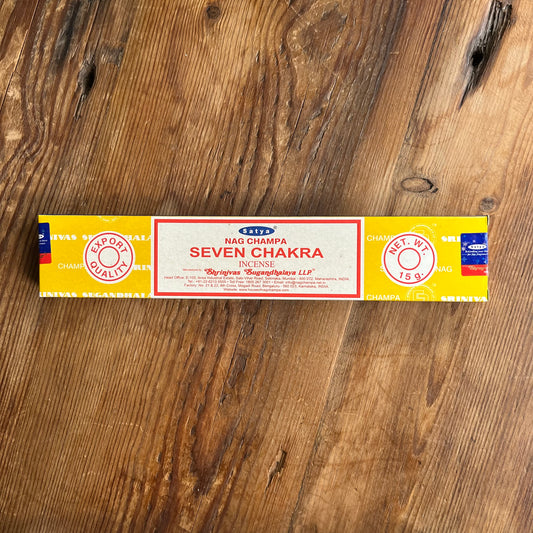 A rectangular box of Nag CHAMPA SEVEN CHAKRA INCENSE sticks lies on a rustic wooden surface. The packaging is primarily yellow with a central cream-colored label bordered in red, featuring blue and red brand details.