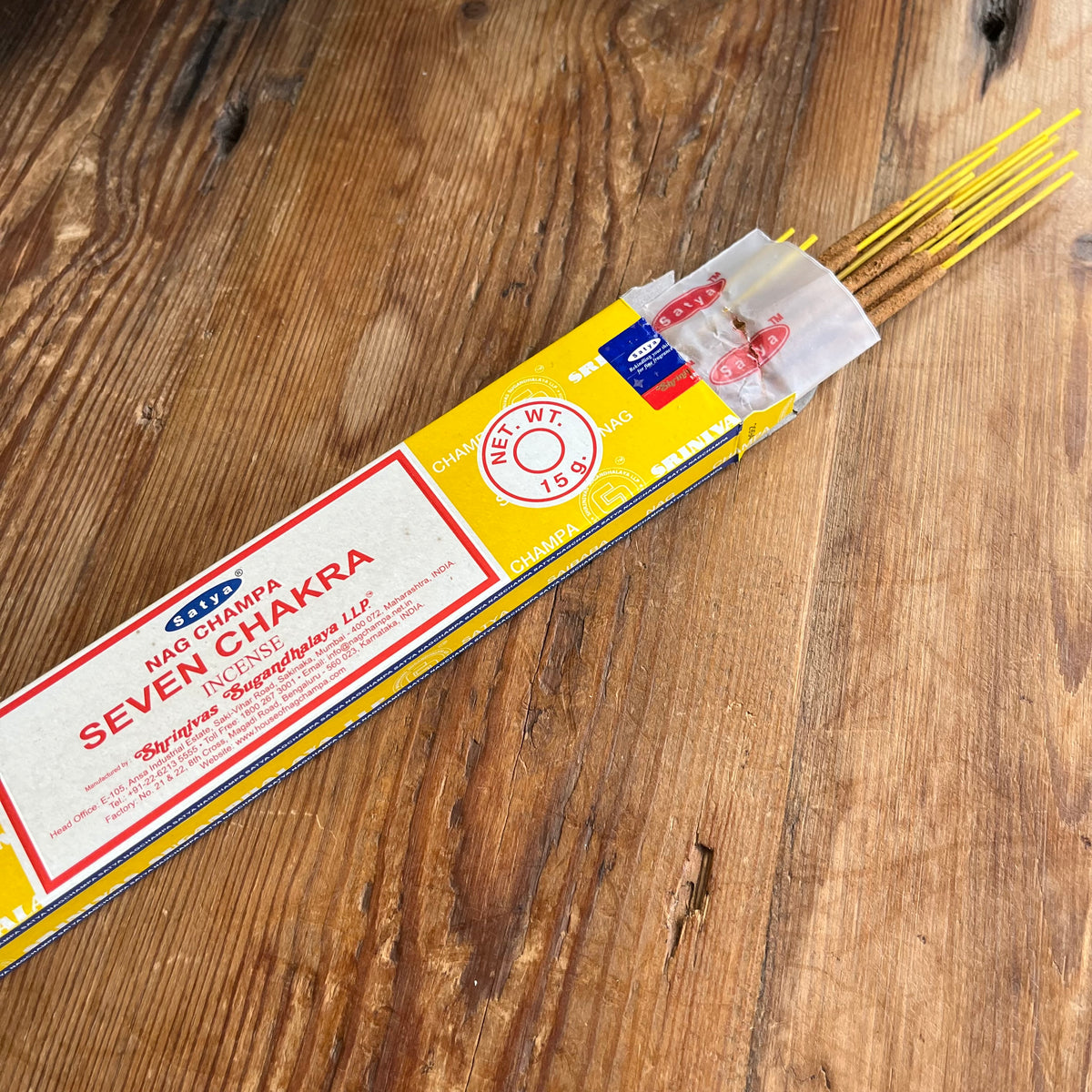 A package of Nag Champa Seven Chakra incense sticks placed diagonally on a rustic wooden surface. The packaging is bright yellow and white with red text, and several incense sticks are visible extending from the top right.