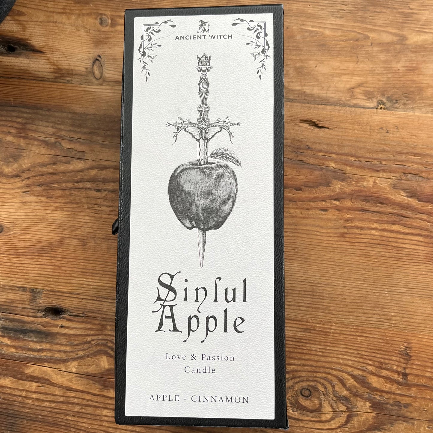 A tall rectangular candle box labeled 'Ancient Witch Sinful Apple Love & Passion Candle' with an illustration of a dagger piercing an apple, placed on a rustic wooden surface.
