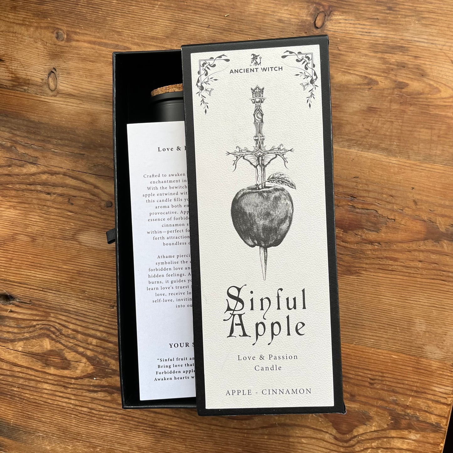 An open black gift box containing a candle and its front label, placed on a wooden tabletop. The label reads 'Ancient Witch' and 'Sinful Apple' with an illustration of a dagger piercing an apple.