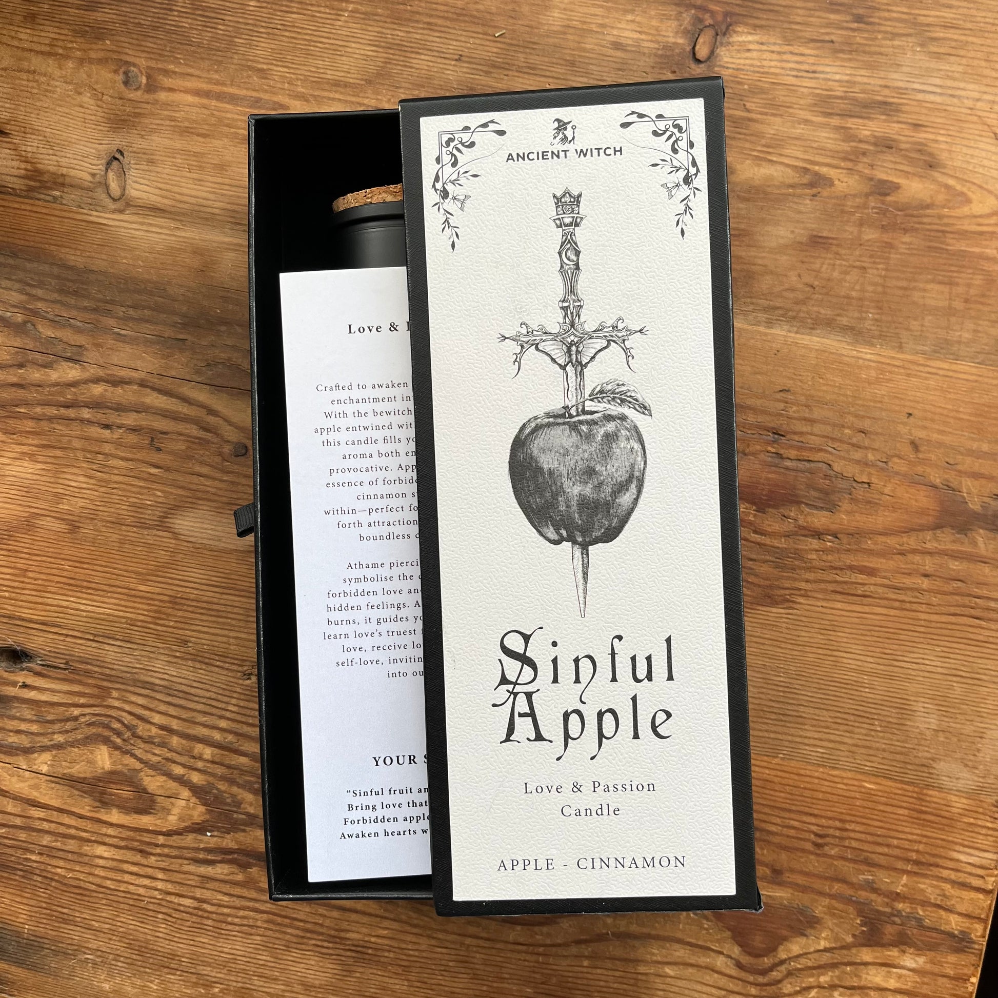 An open black gift box containing a candle and its front label, placed on a wooden tabletop. The label reads 'Ancient Witch' and 'Sinful Apple' with an illustration of a dagger piercing an apple.