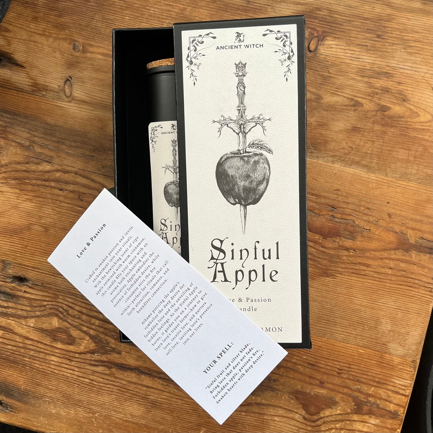 A boxed candle set on a wooden tabletop, featuring a spell card with a black illustration of a dagger through an apple and the text 'Ancient Witch Sinful Apple,' a tall candle in a glass holder and a presentation box.