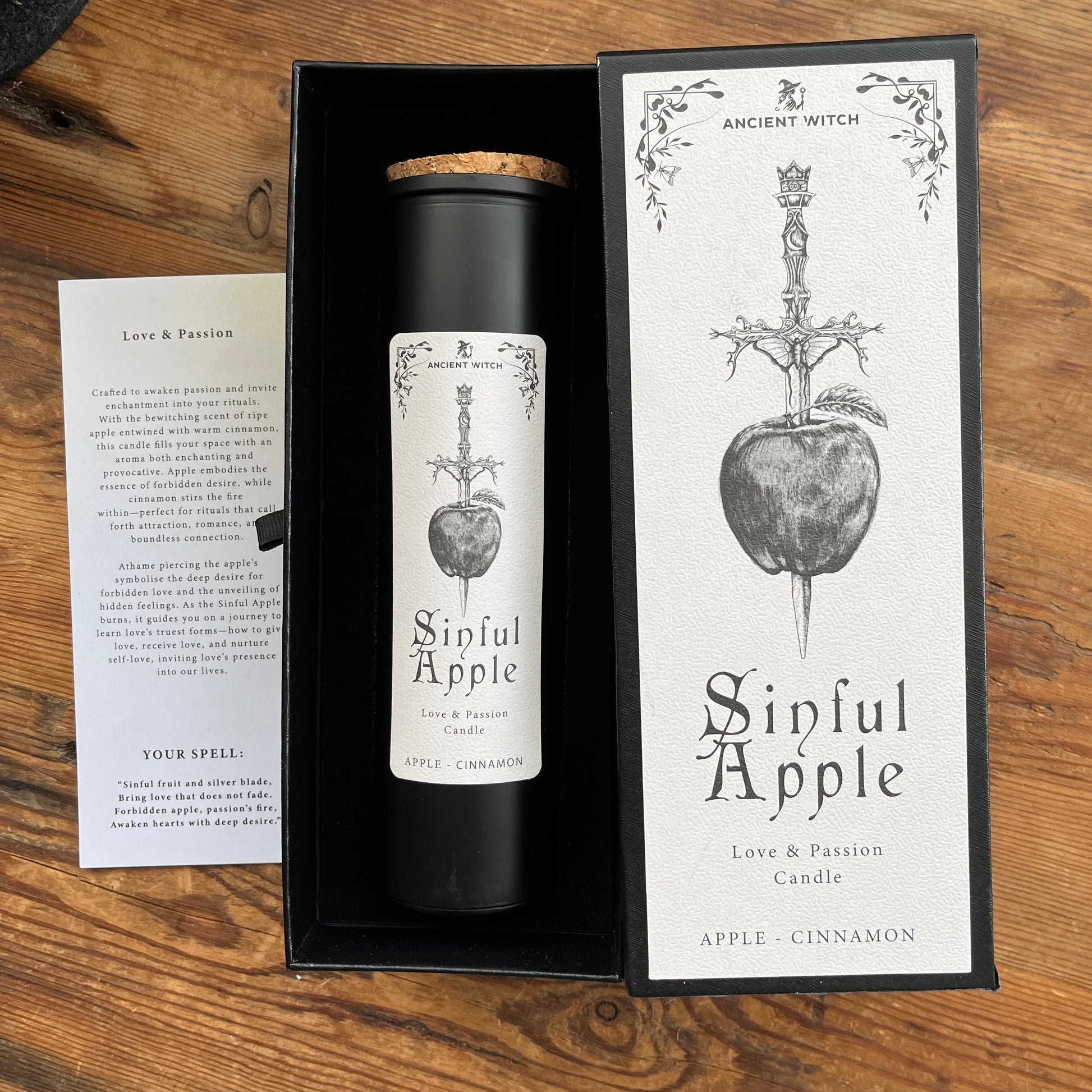 A black cylindrical candle in a black gift box with its matching printed outer box beside it, plus a small printed card on a wooden surface. The candle label and packaging both show the brand name Ancient Witch and the product name Sinful Apple, with an apple-and-sword illustration.