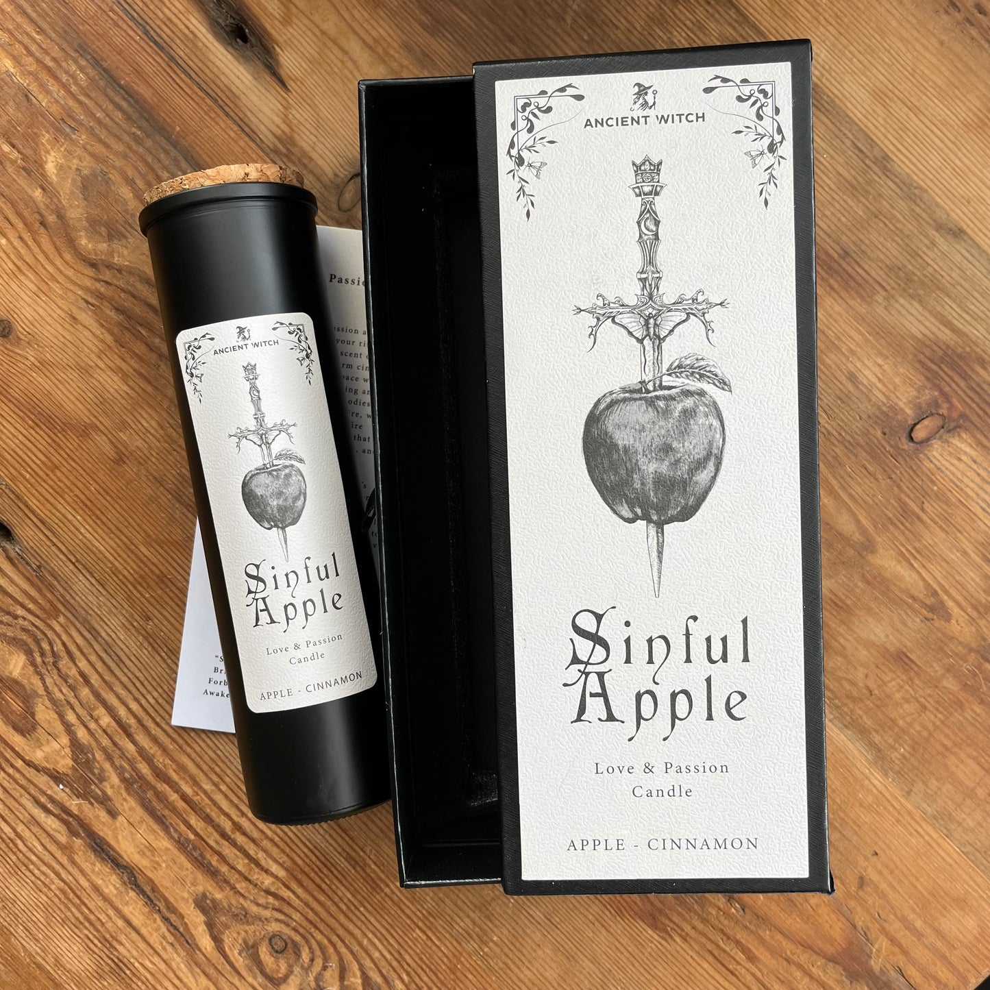 A black cylindrical candle jar with a cork stopper and a white label reading 'Sinful Apple' sits beside its matching black-and-white product box on a wooden surface. The label and box feature an illustration of a dagger piercing an apple, with the brand name 'Ancient Witch' visible.