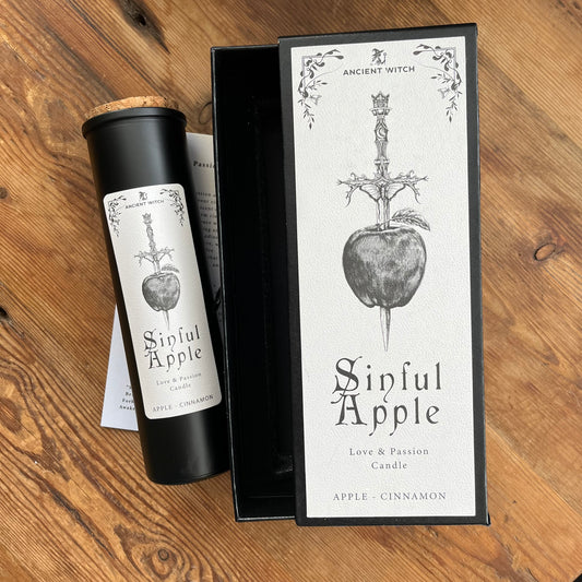 A black cylindrical candle jar with a cork stopper and a white label reading 'Sinful Apple' sits beside its matching black-and-white product box on a wooden surface. The label and box feature an illustration of a dagger piercing an apple, with the brand name 'Ancient Witch' visible.