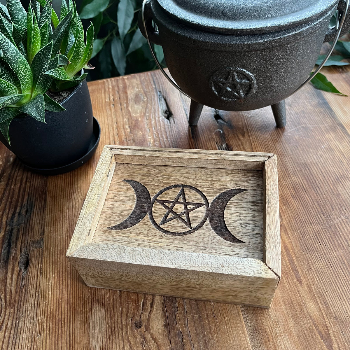 A rectangular wooden box with a laser-engraved triple moon and pentagram design on the lid, displayed on a rustic wooden table next to a potted succulent and a black cast iron cauldron.