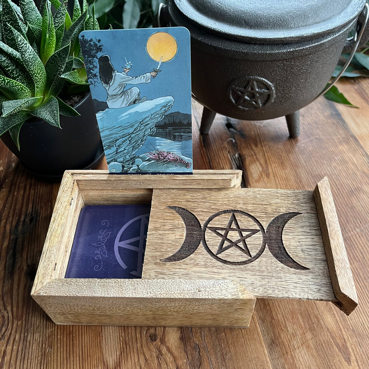 A wooden box with a sliding drawer containing tarot cards, one of which is pulled out. The box is engraved with triple moon and pentagram symbols.