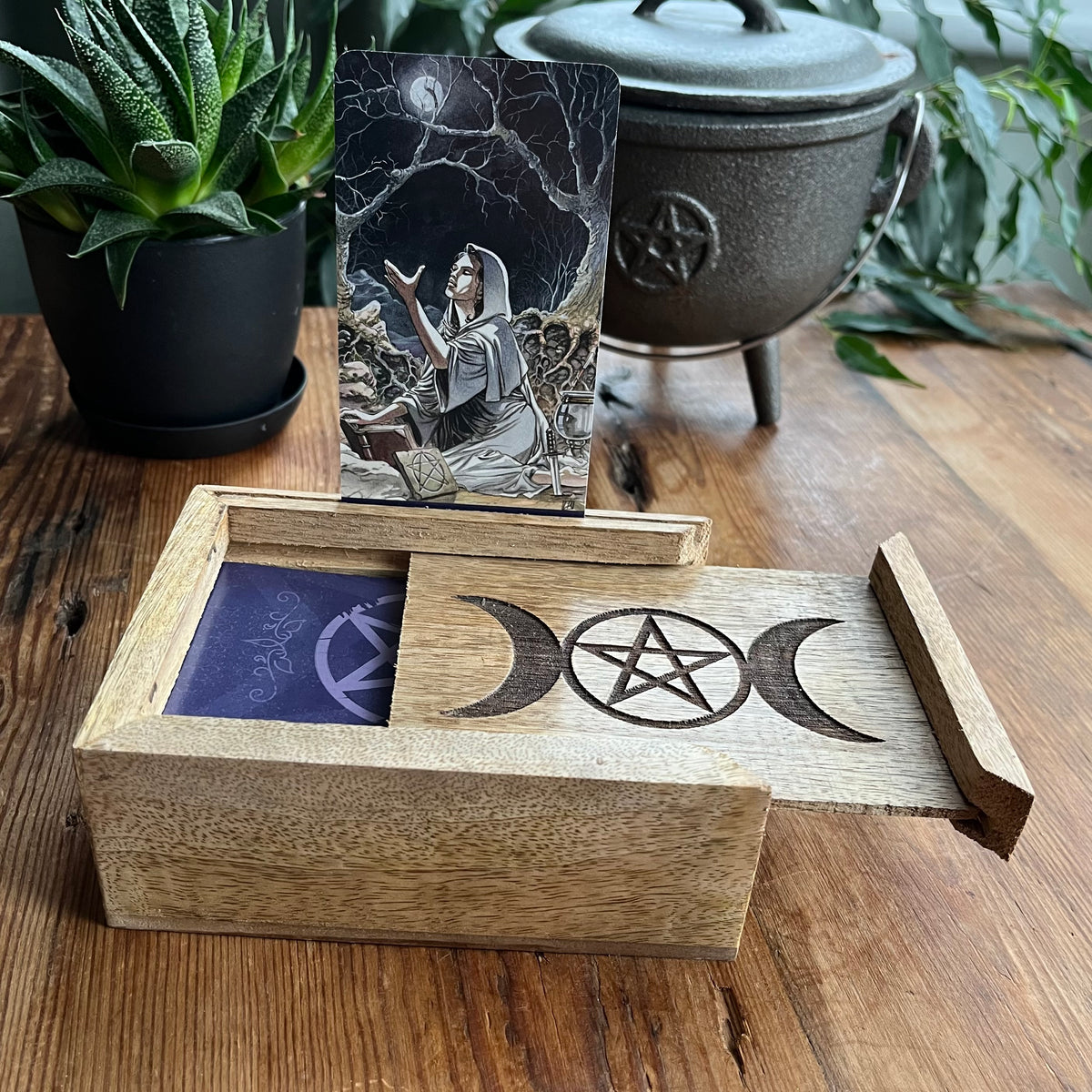 A wooden tarot card box with engraved moon and pentagram symbols, shown open with a tarot card standing inside and a purple card sleeve visible. The box sits on a wooden table with potted plants and a cast iron pot in the background.