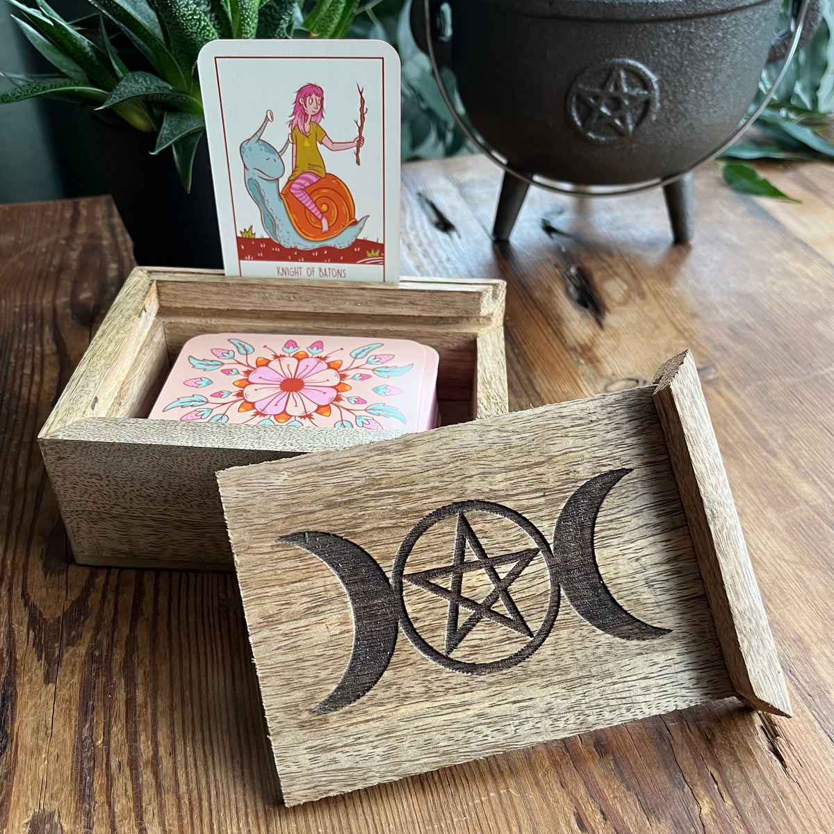 A wooden box with a carved triple moon and pentagram design on the lid, holding a tarot card labeled 'Knight of Wands' on a wooden tabletop with a plant and cauldron in the background.