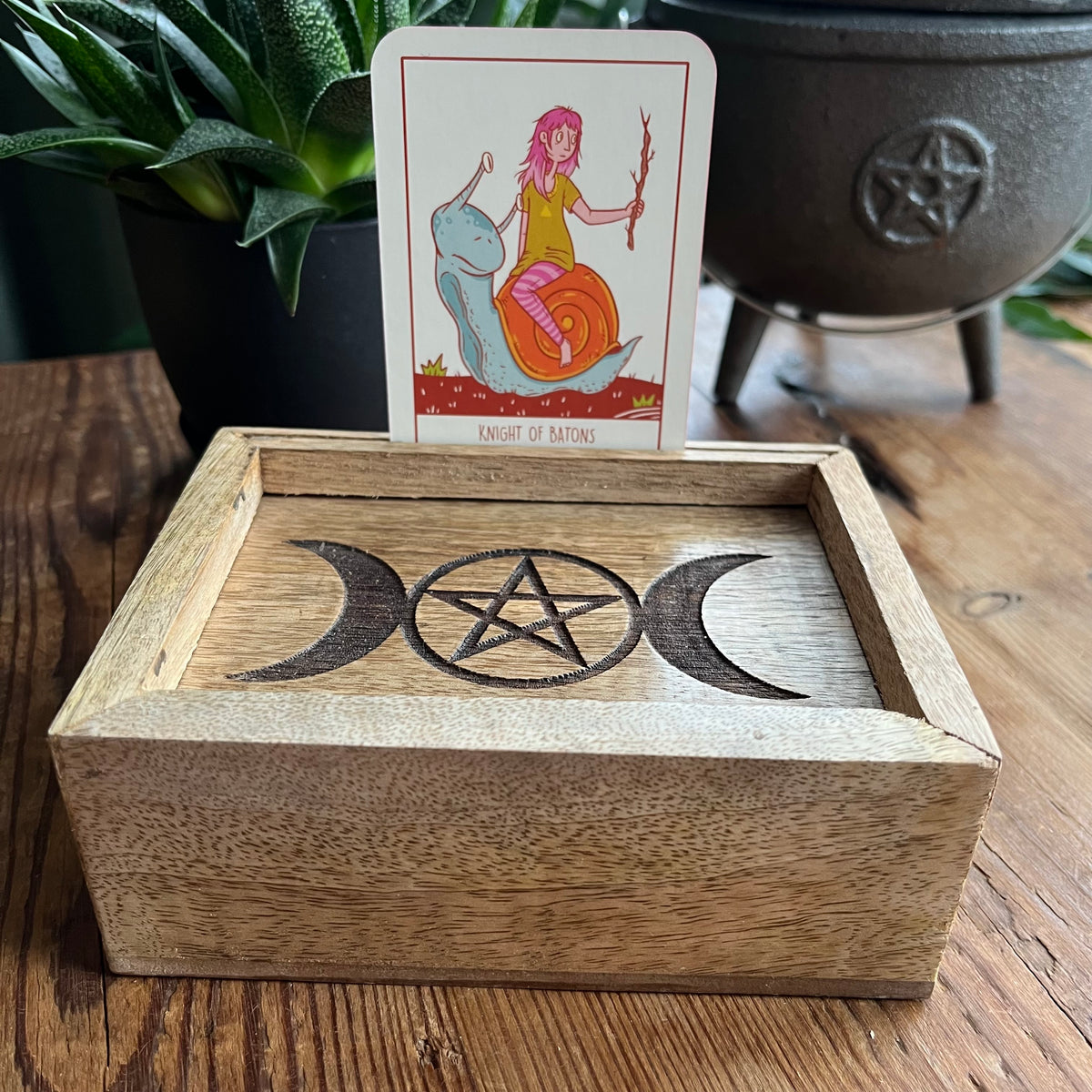 A wooden box with a carved triple moon and pentagram design on the lid, holding a tarot card labeled 'Knight of Wands' on a wooden tabletop with a plant and cauldron in the background.