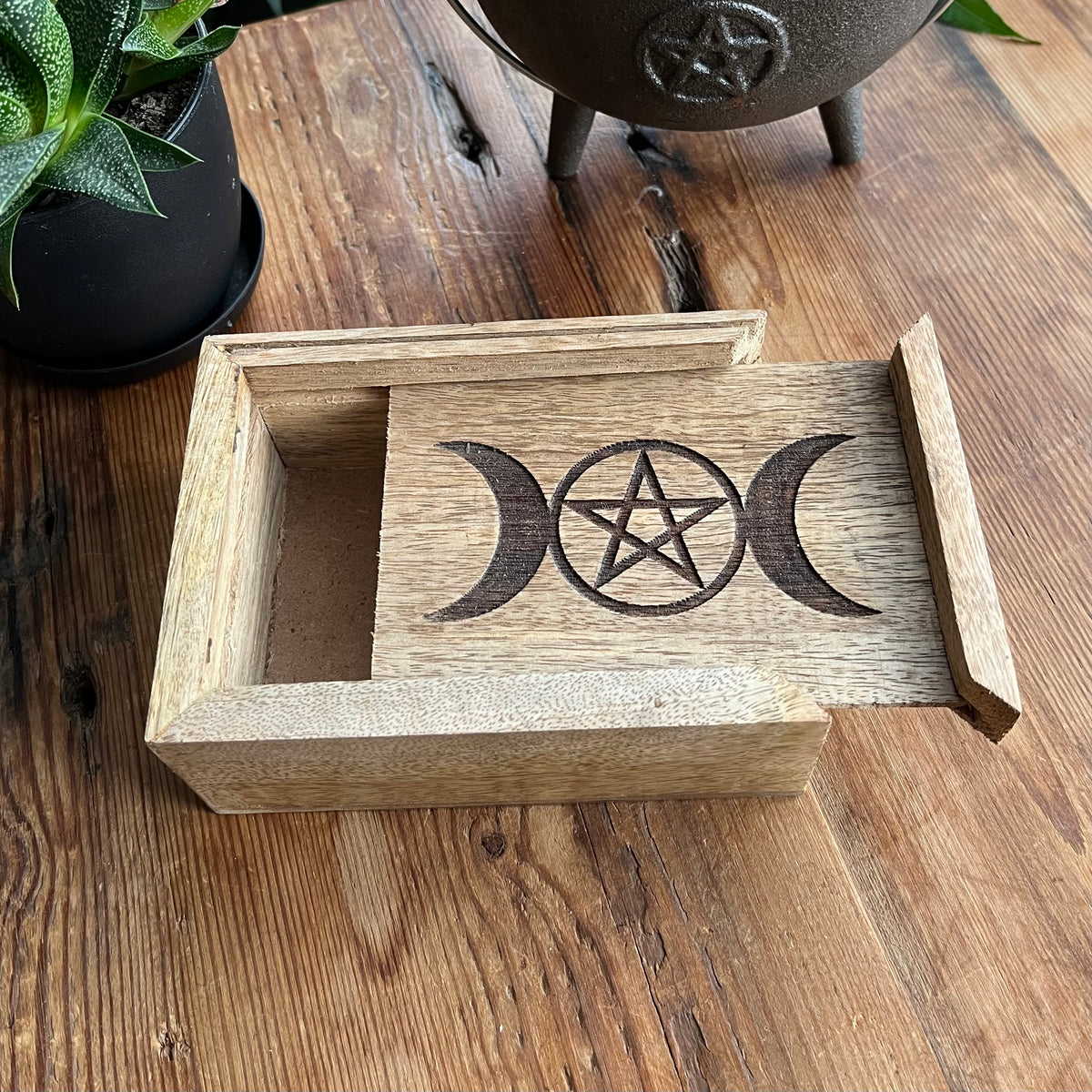 A rectangular wooden box with a laser-engraved triple moon and pentagram design on a sliding lid, displayed on a rustic wooden table next to a potted succulent and a black cast iron cauldron.