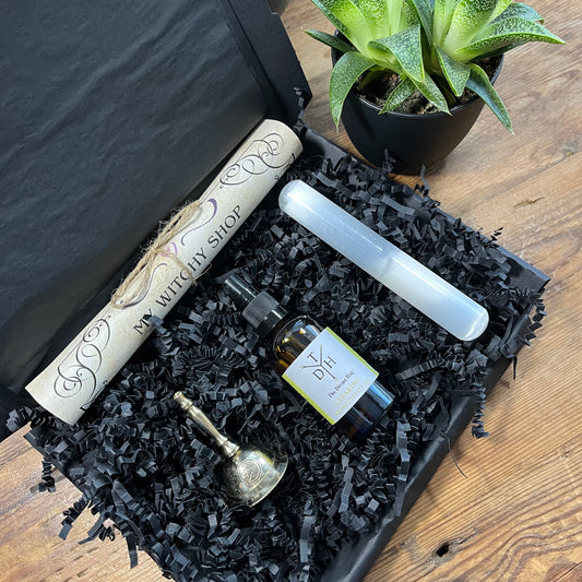 Gift box with 'My Witchy Shop' scroll, bottle of energy clearing room spray, Selenite wand and bell on black crinkled paper.